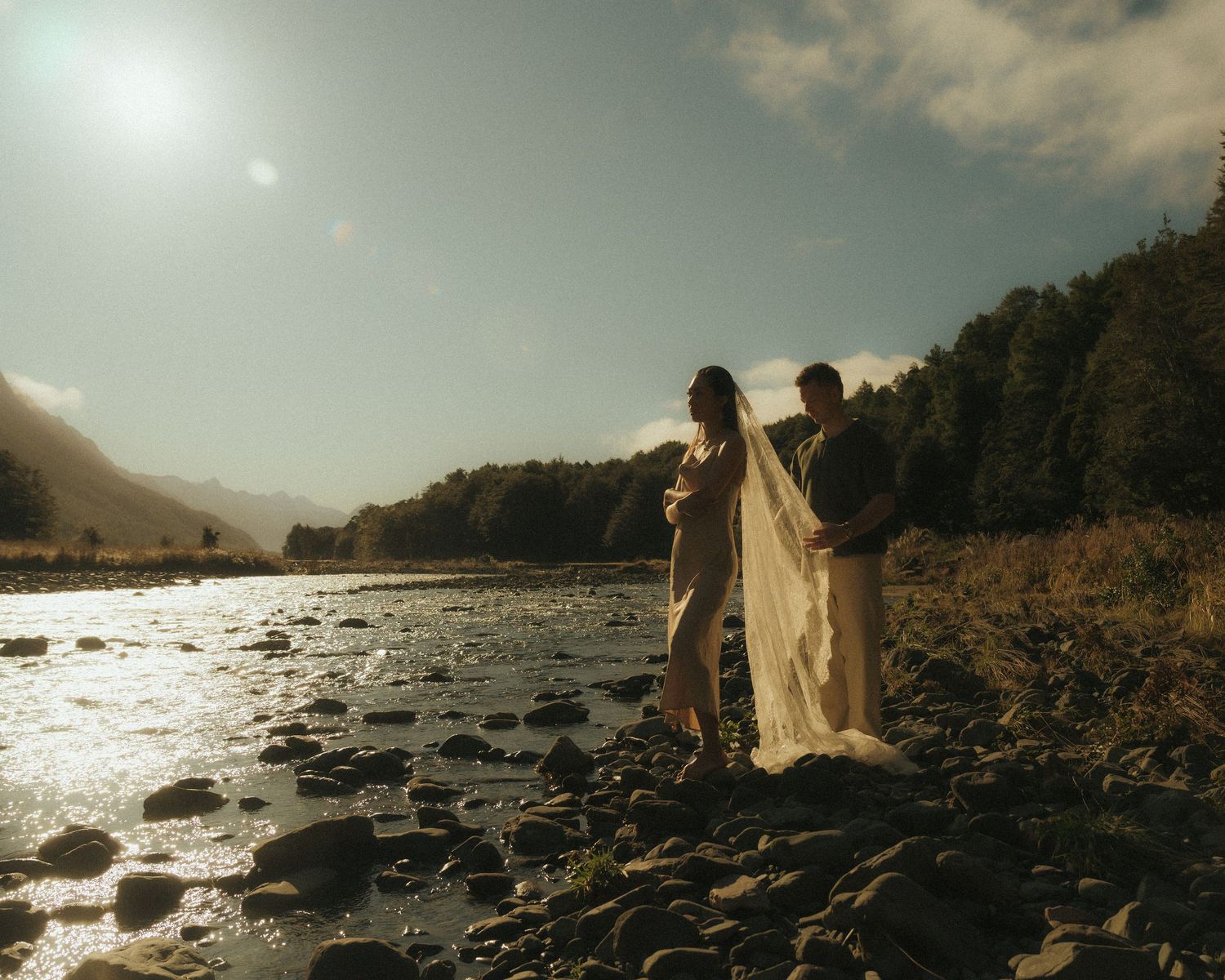 Couple in wedding attire standing on rocky riverbank with mountains and glowing sunlight in background.