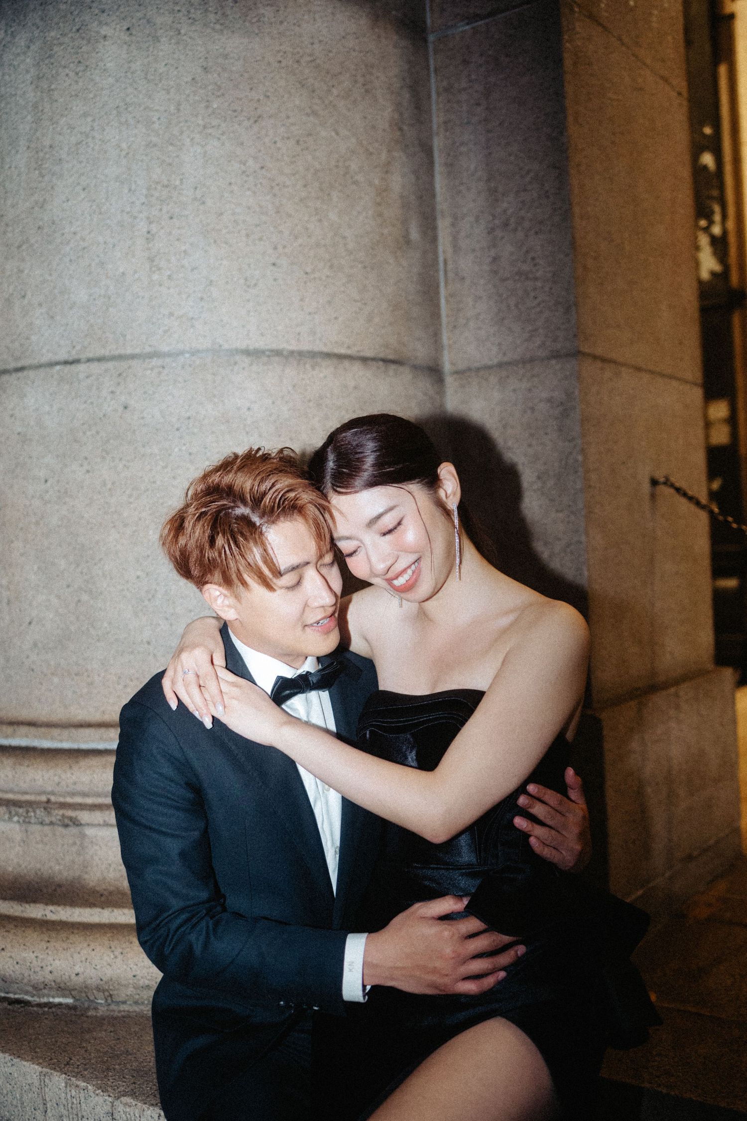 A couple in elegant formal wear shares a tender embrace against stone columns at night.
