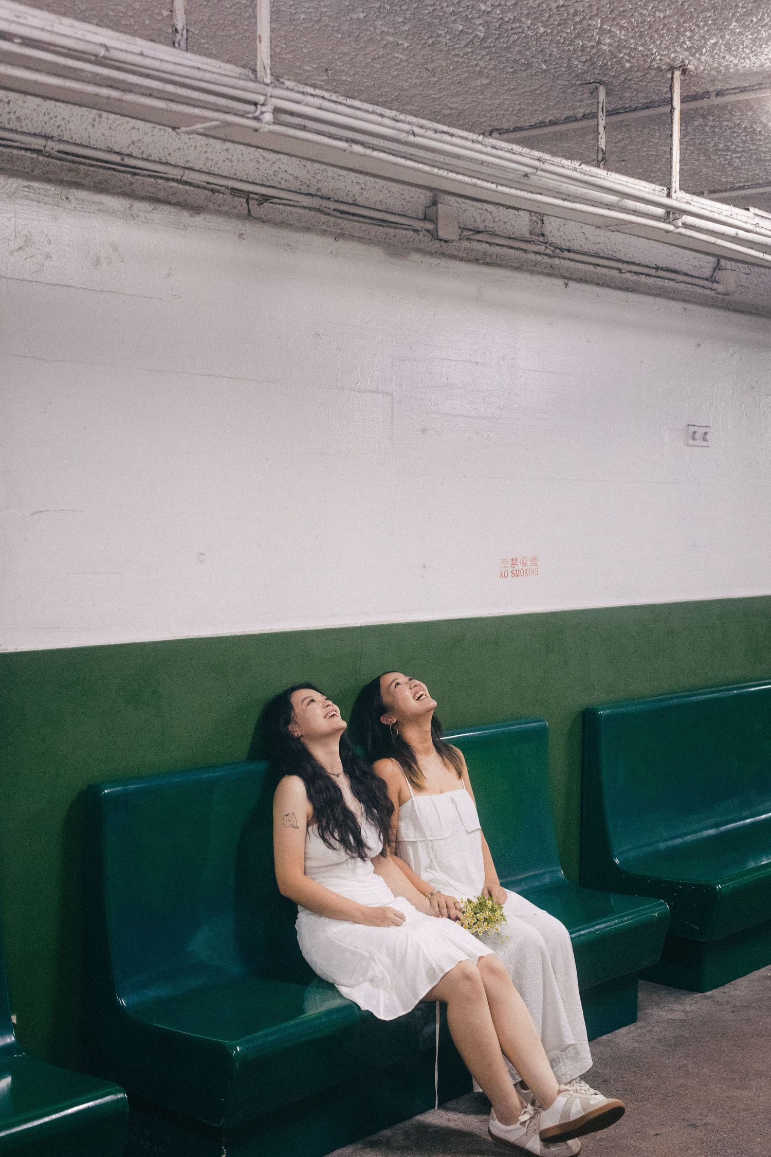 Two young Asian women in white dresses sitting on green benches, heads tilted back.