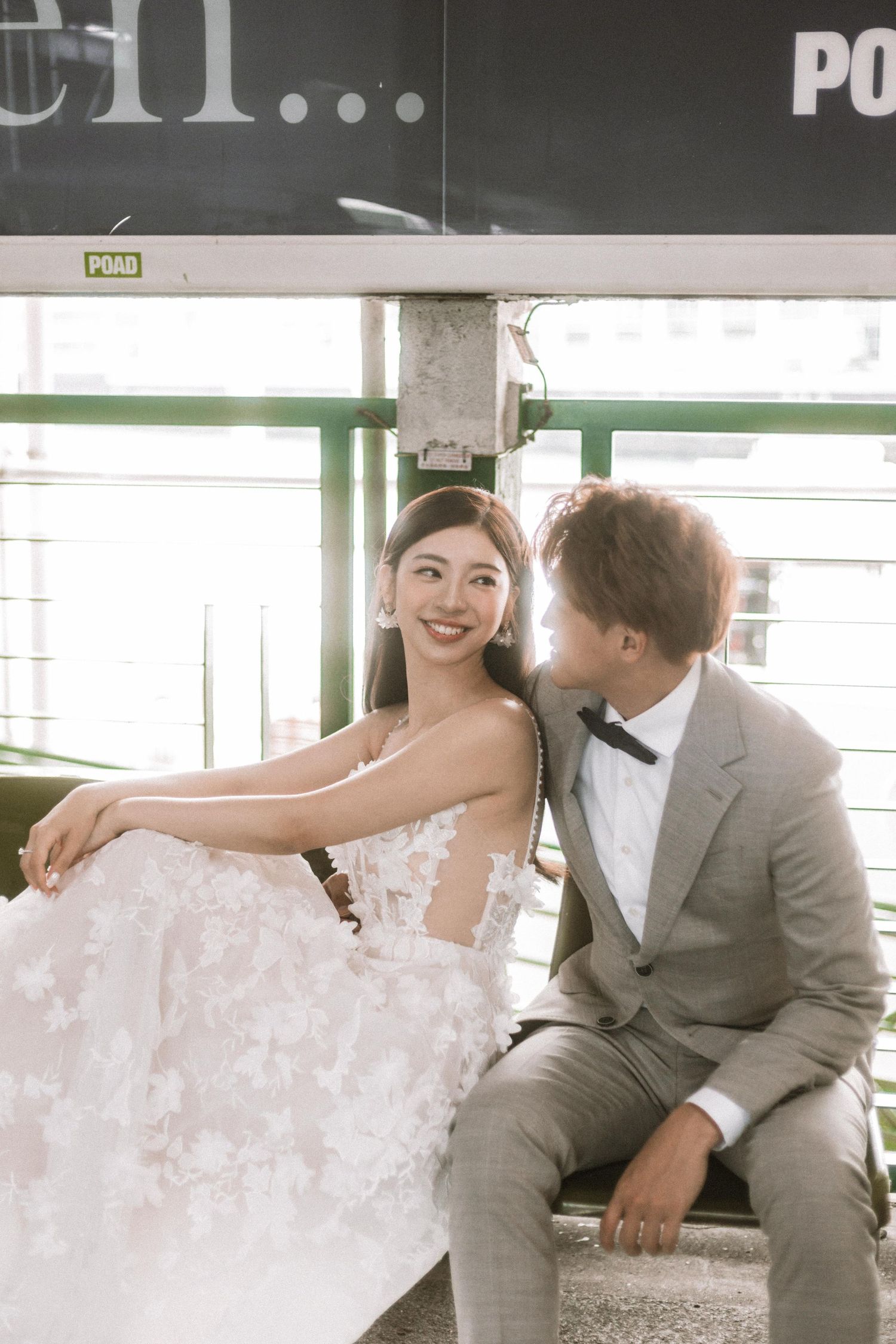 A happy couple in wedding attire sits together on a tram bench, bride in floral white gown and groom in gray suit with bow tie.