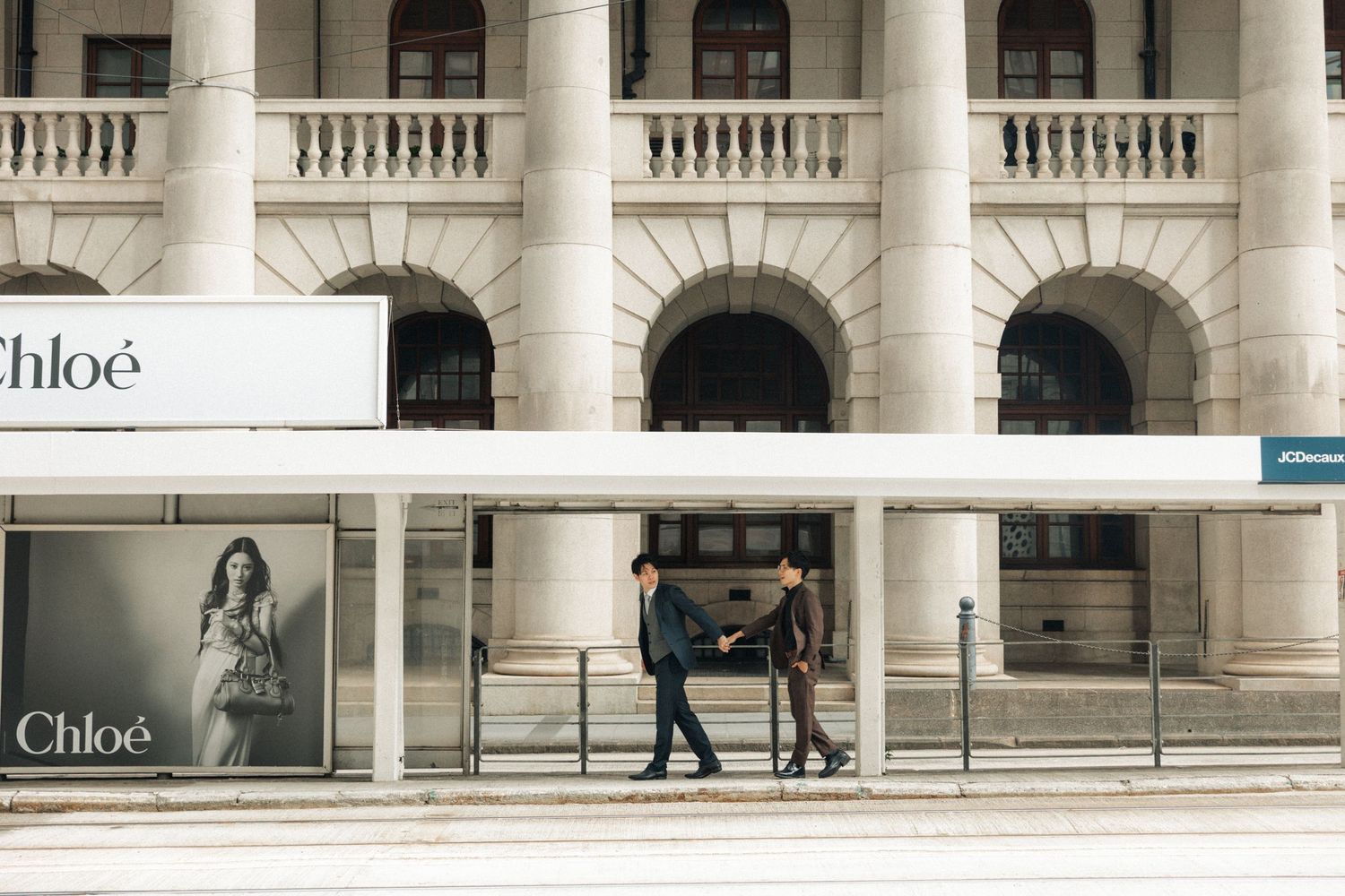 Two men in suits shaking hands outside a grand neoclassical building with Chloe storefront displays.