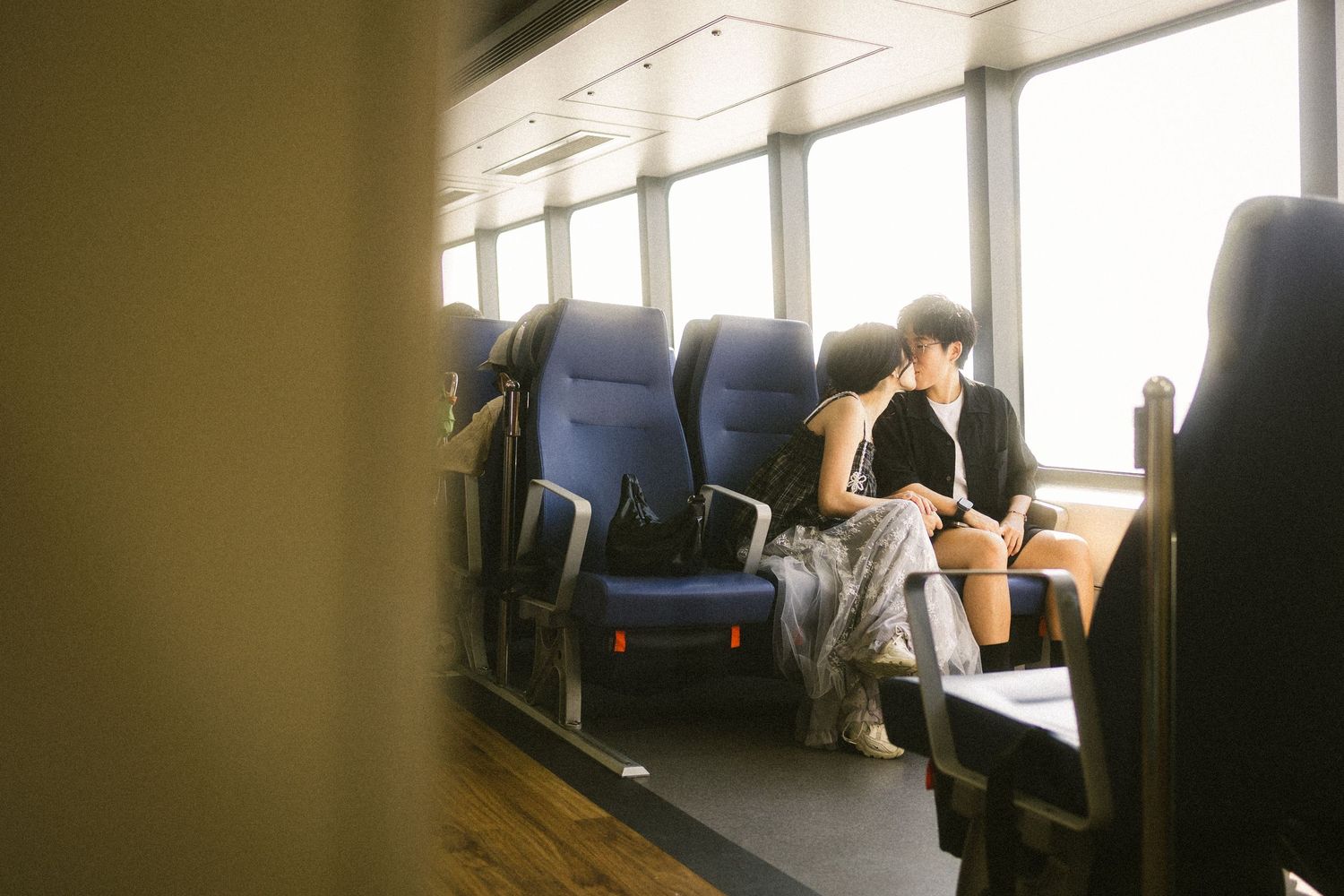 Young couple sitting close together in blue ferry seats bathed in warm sunlight.