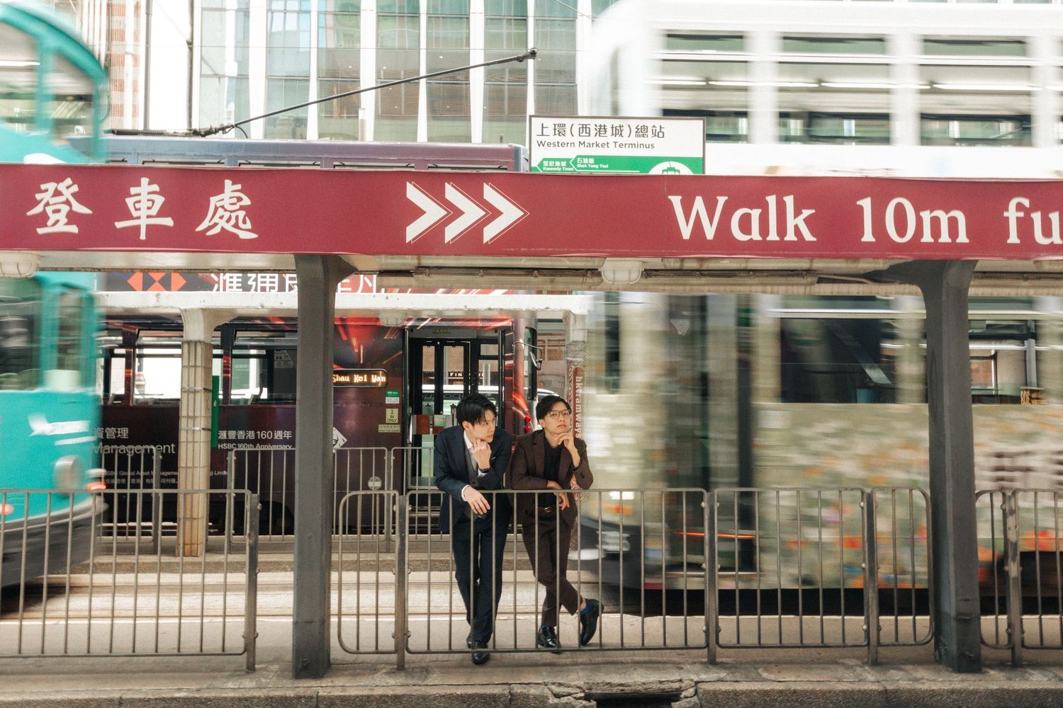 Two people walk under a red Hong Kong tram stop sign as a tram speeds past.