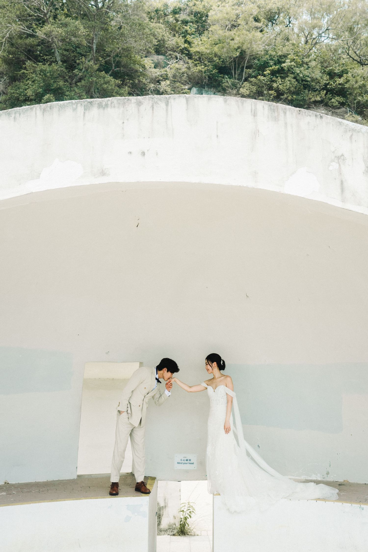 Wedding couple shares intimate moment under white arched concrete structure with lush greenery backdrop.