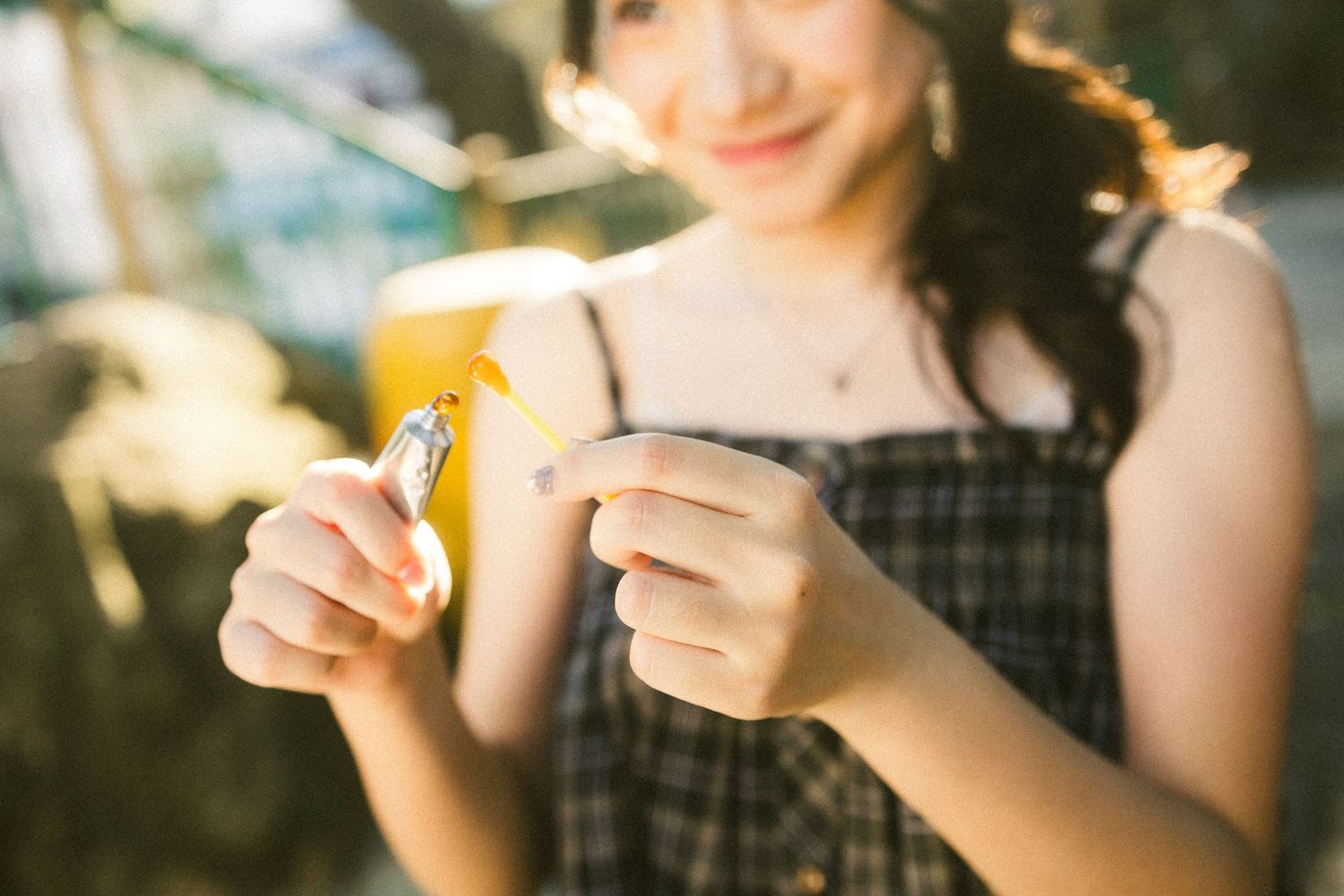 Smiling young woman in plaid dress holding a small object near a yellow drink in sunlight.