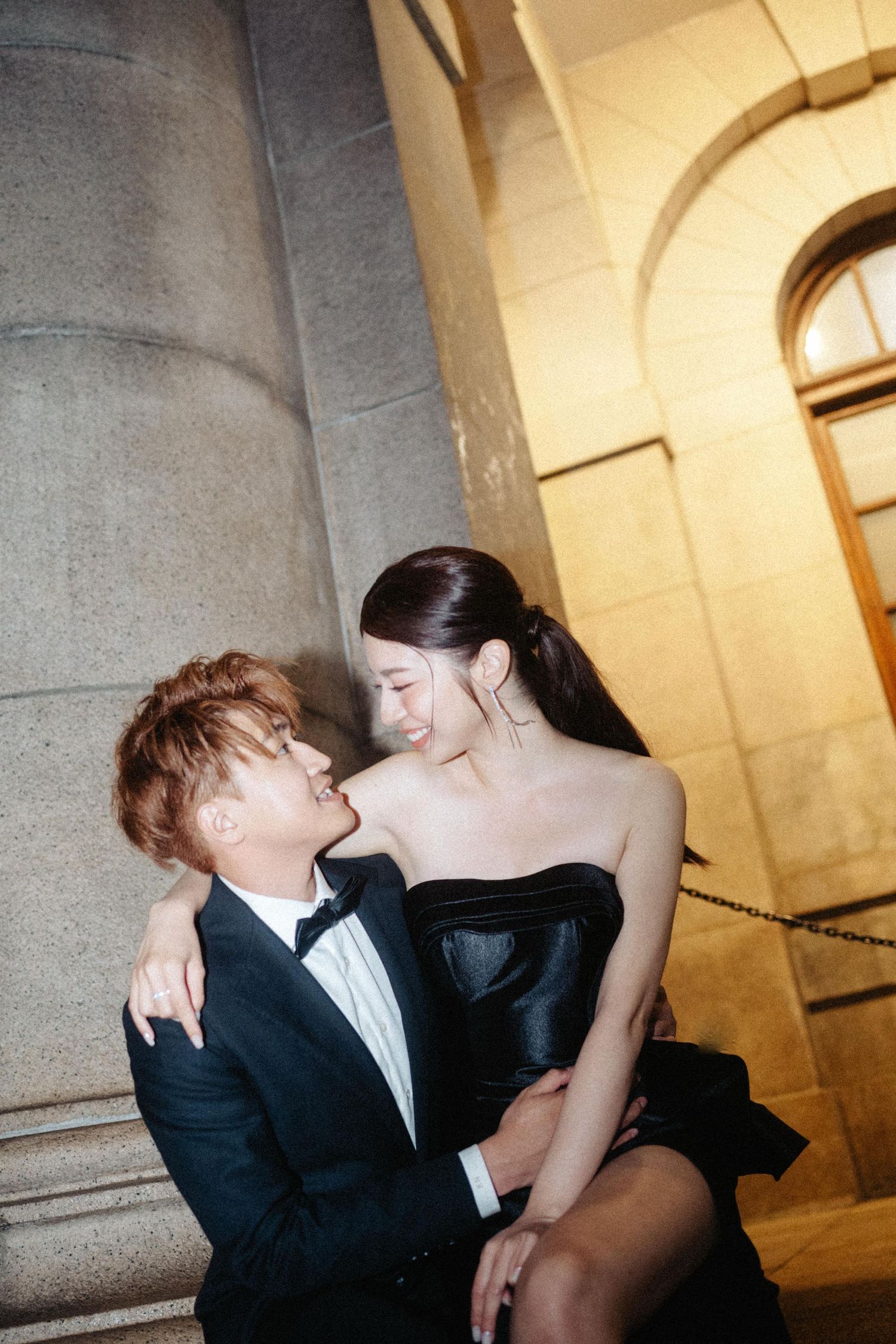 A romantic couple in black formal wear gazes at each other while seated on stone steps of a grand building at night.
