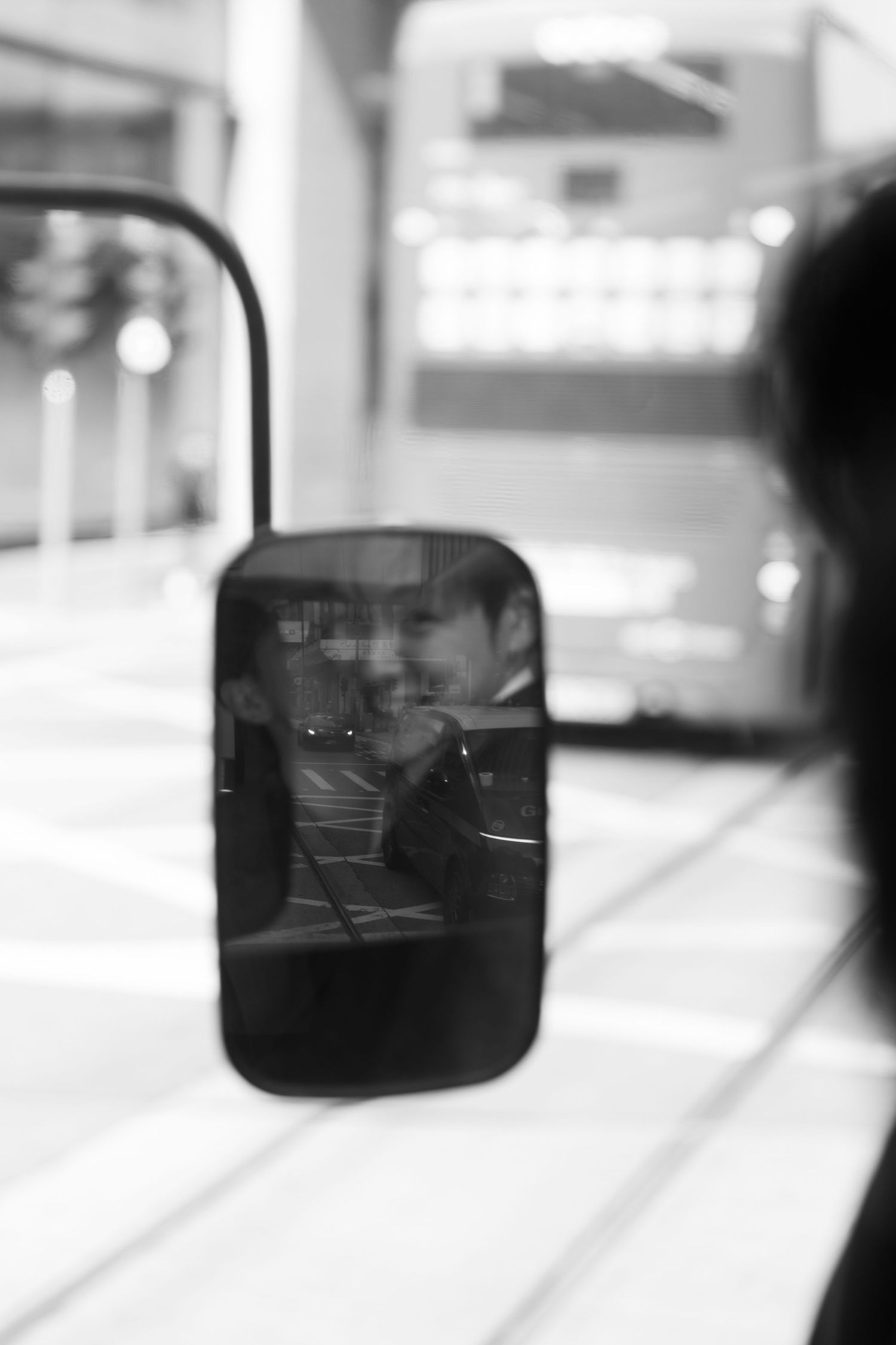 Black and white reflection of a smiling man captured in a tram side mirror on a city street.