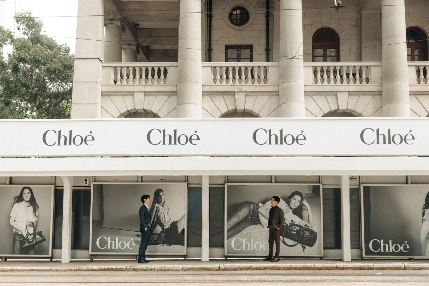 Two men in suits walking past elegant Chloe fashion store windows on a neoclassical building facade.