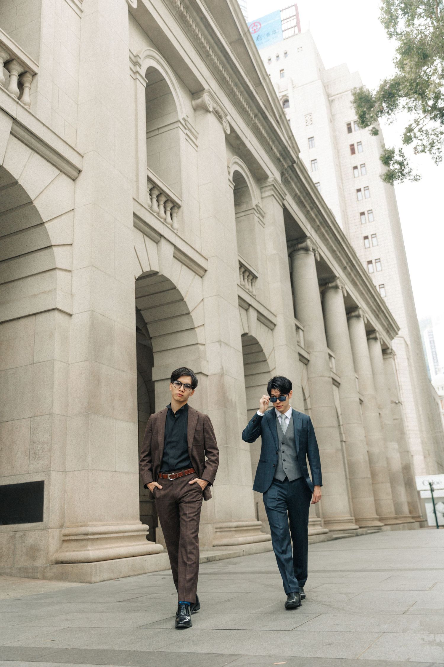 Two Asian men in formal suits walk past a grand colonial building with stone arches.