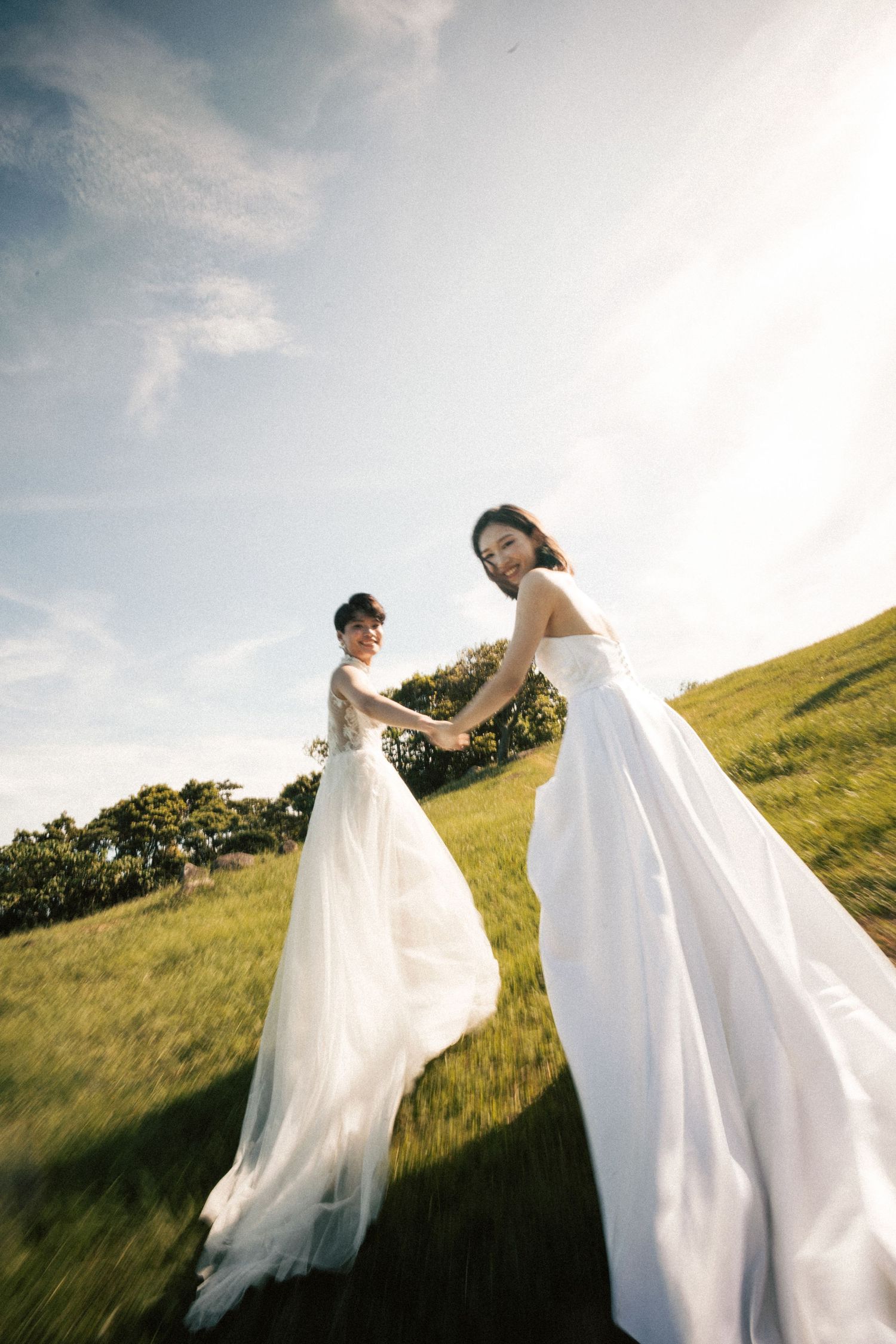 Two brides in white gowns hold hands while walking through a sunlit green field under a partly cloudy sky.