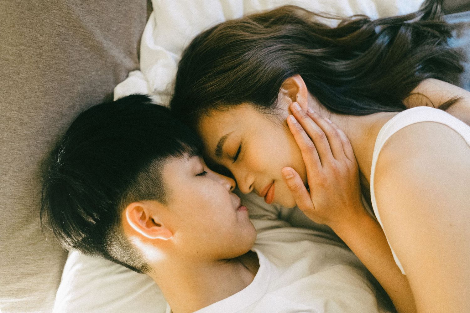 A young Asian couple lying close together on white pillows, sharing a tender intimate moment.