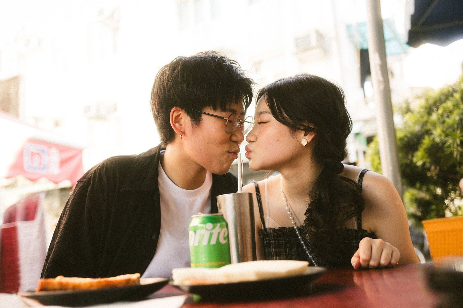Young Asian couple sharing a romantic kiss at an outdoor cafe table with food and drinks.