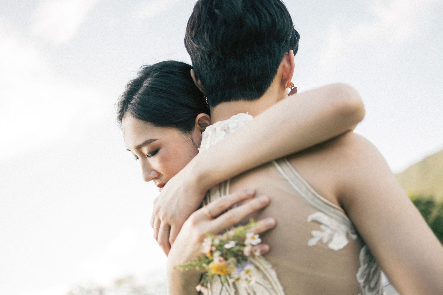 Two brides share a tender embrace outdoors, one holding a wildflower bouquet on their wedding day.