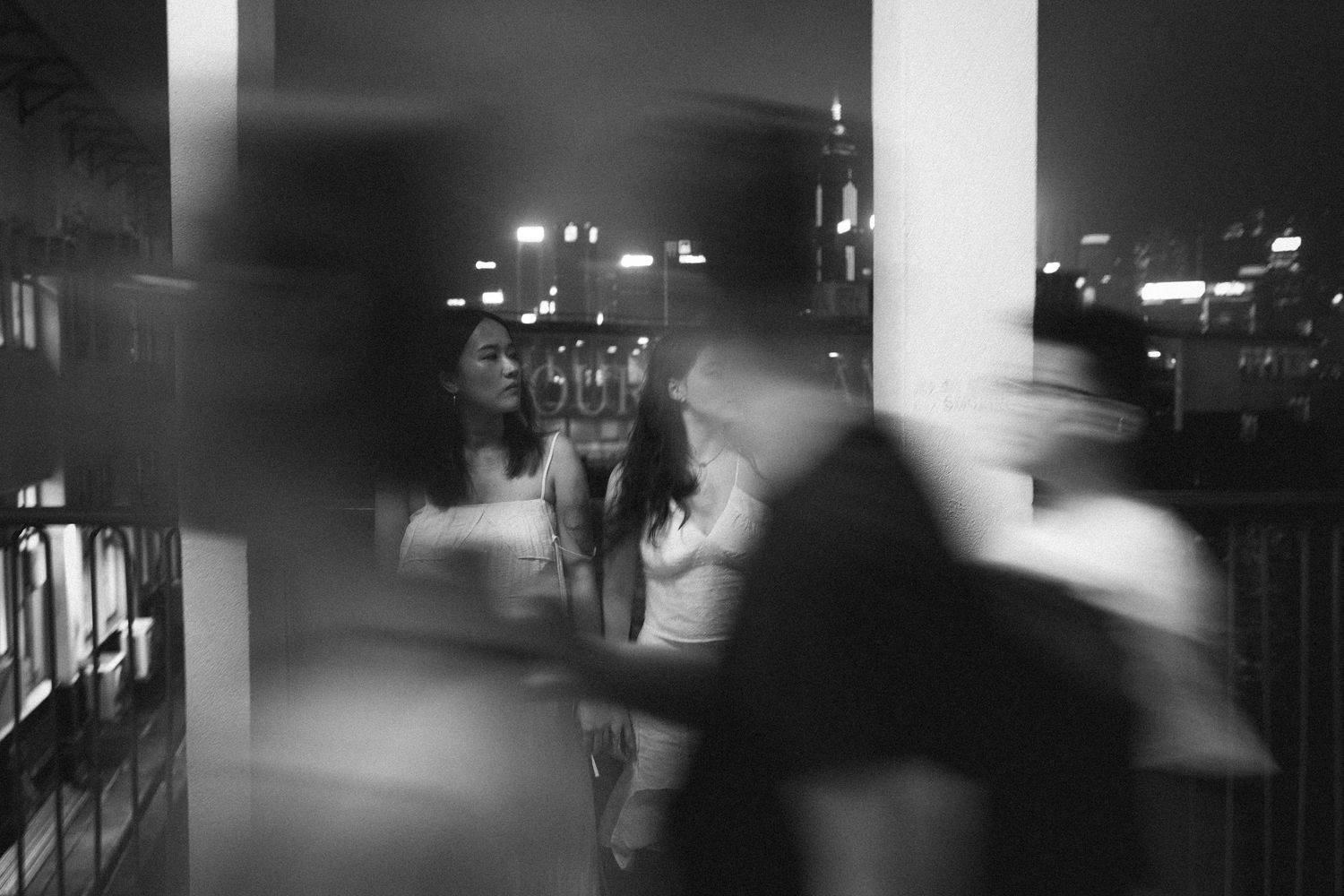 Black and white photo of a woman standing on a balcony at night with blurred figures around her.
