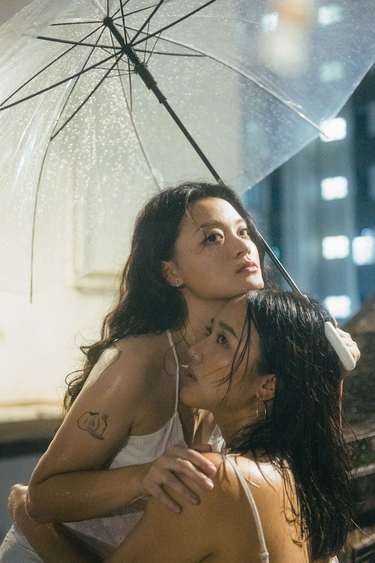 Two women embracing under a clear umbrella on a rainy night, one with tattoos visible.