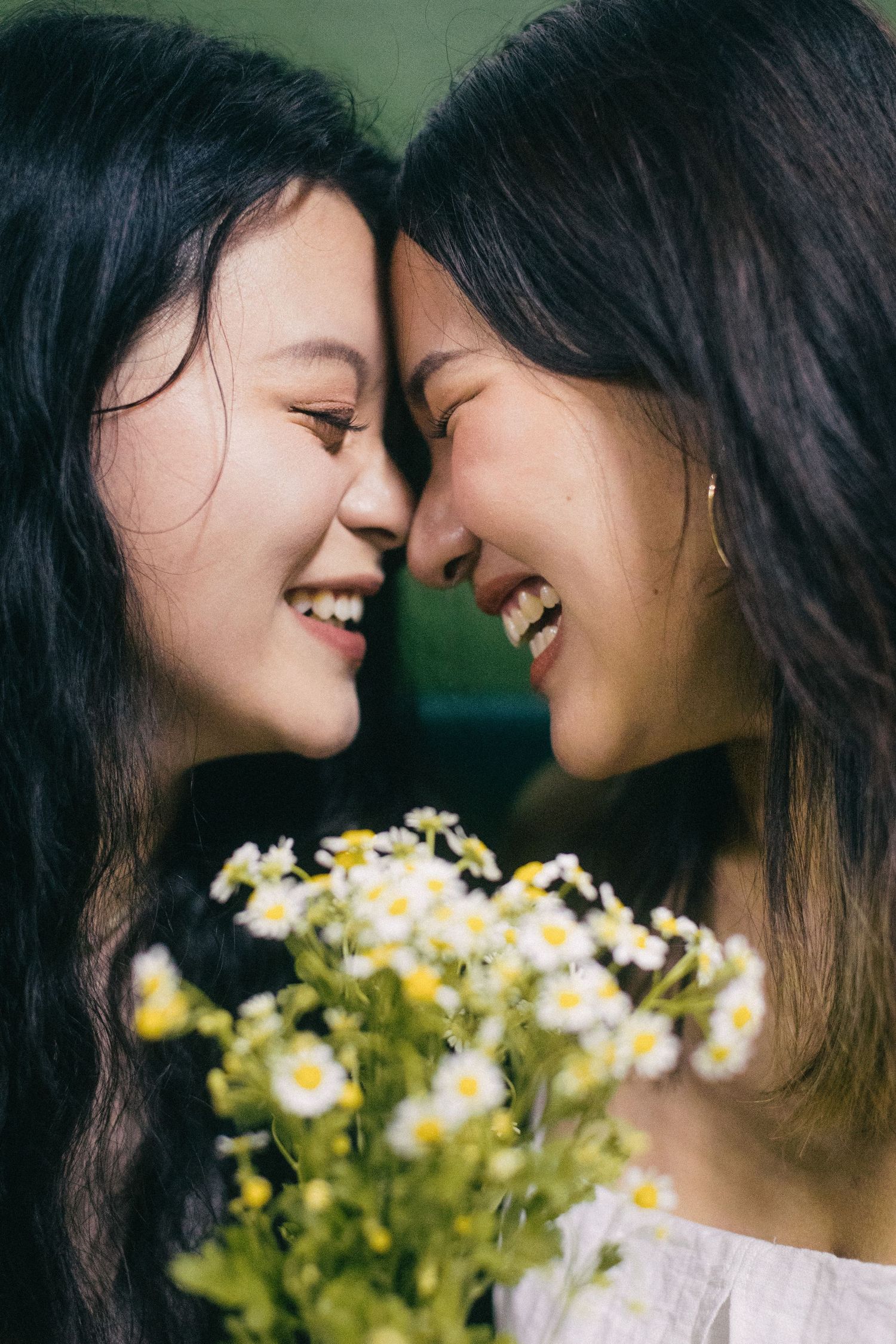 Two smiling Asian women touching foreheads while holding white daisy flowers.