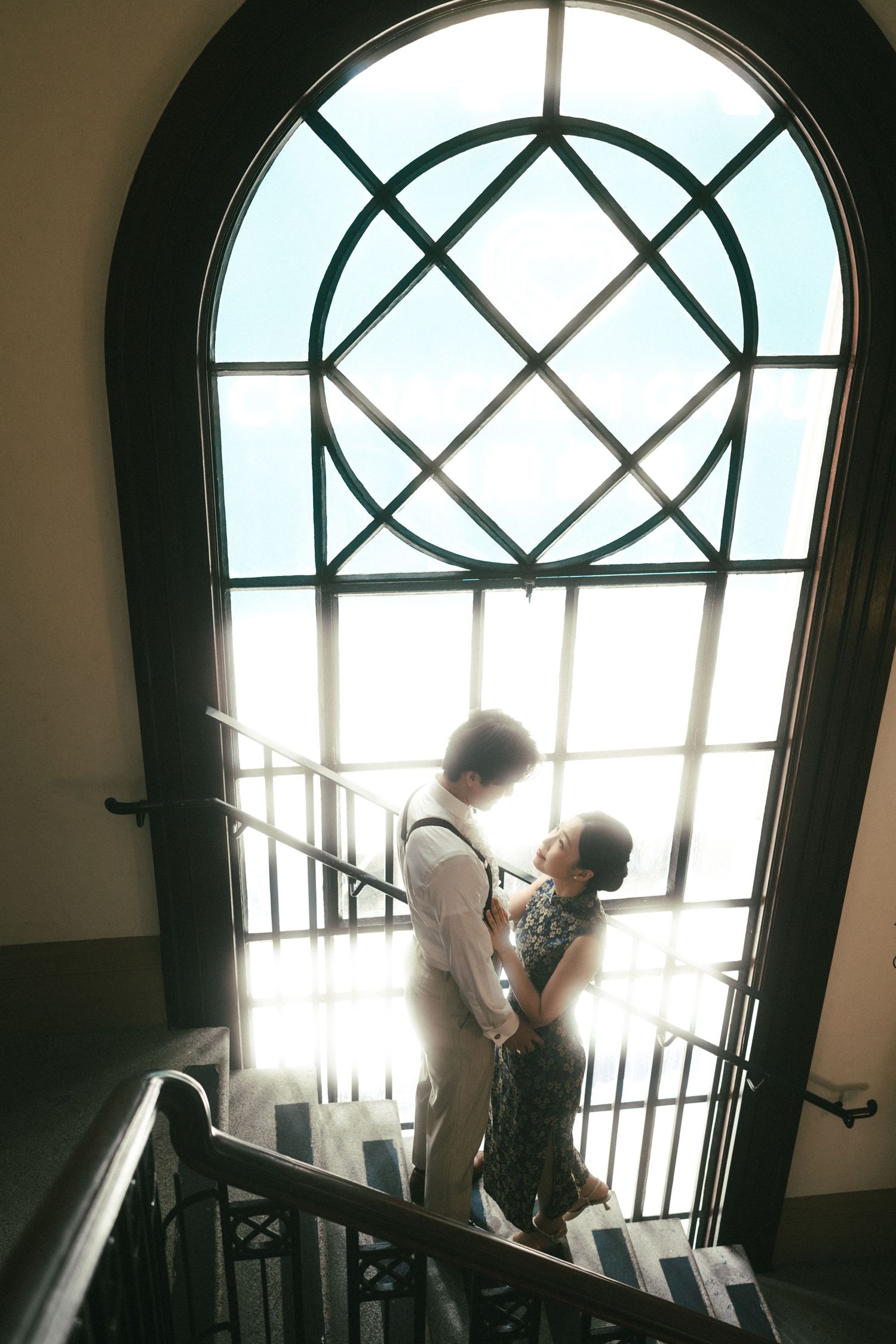 Couple embraces on staircase bathed in sunlight streaming through elegant arched lattice window.