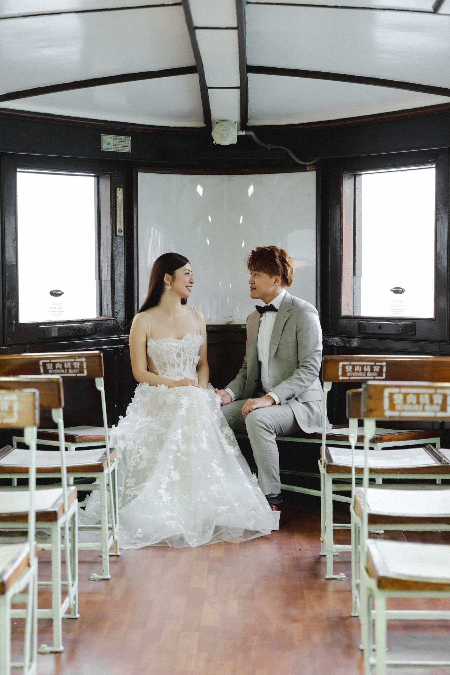 A bride in a white gown and groom in a gray suit hold hands inside a vintage tram.