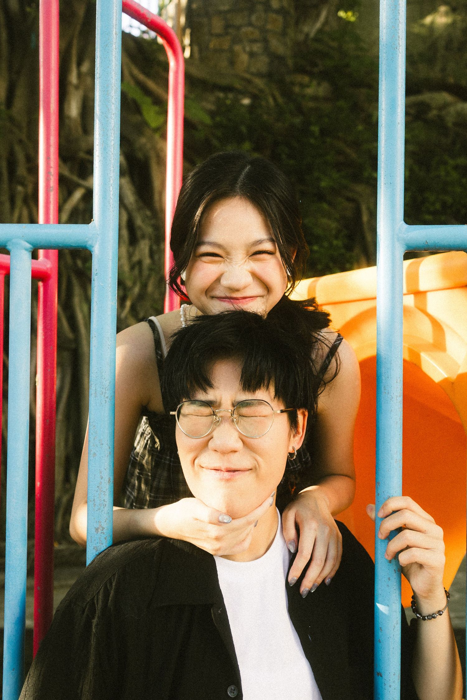 Smiling young woman rests her chin on a friend's head near colorful playground equipment.