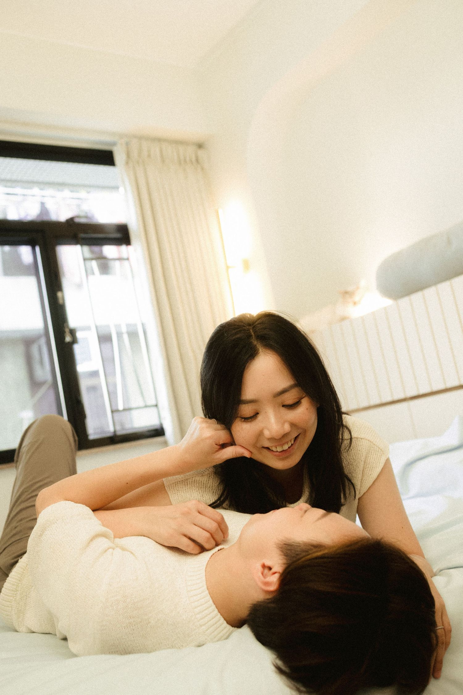 A smiling Asian woman lying on a bed looking down at her partner in a bright cozy bedroom.