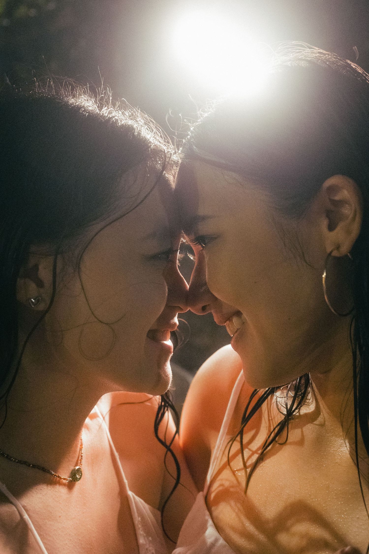 Two women with wet hair touch foreheads tenderly in warm backlit sunlight.