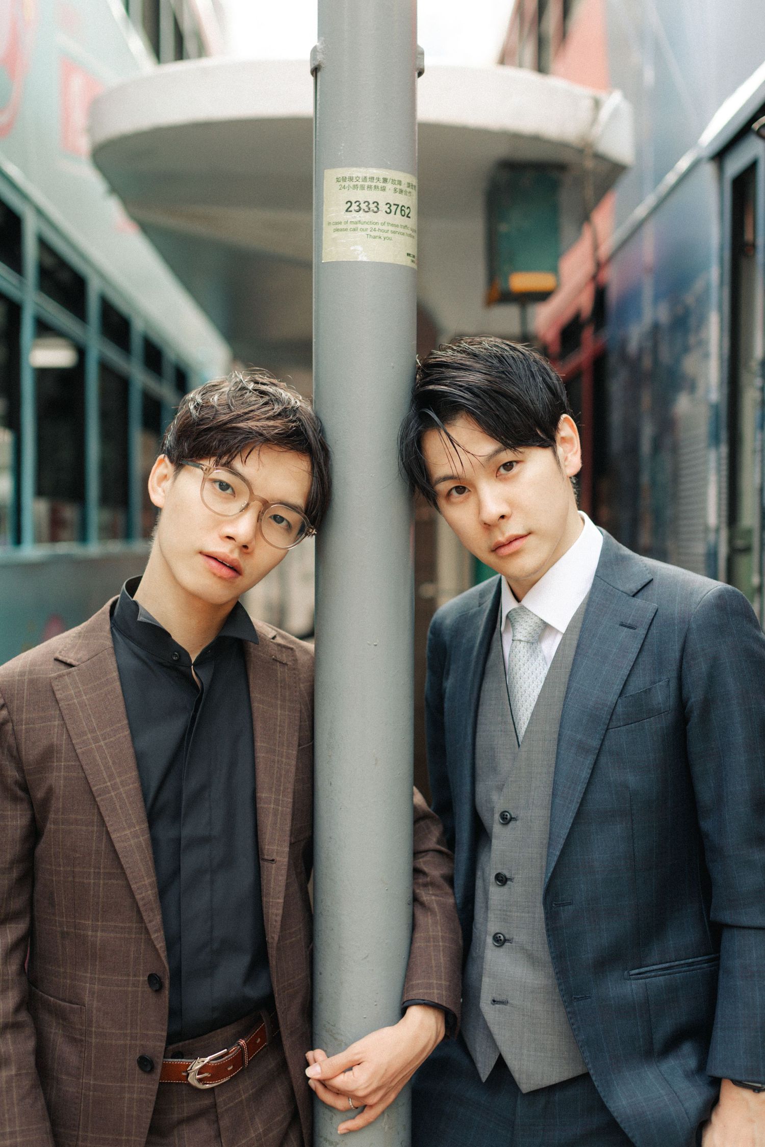 Two stylish Asian men in suits posing beside a street pole in an urban city setting.