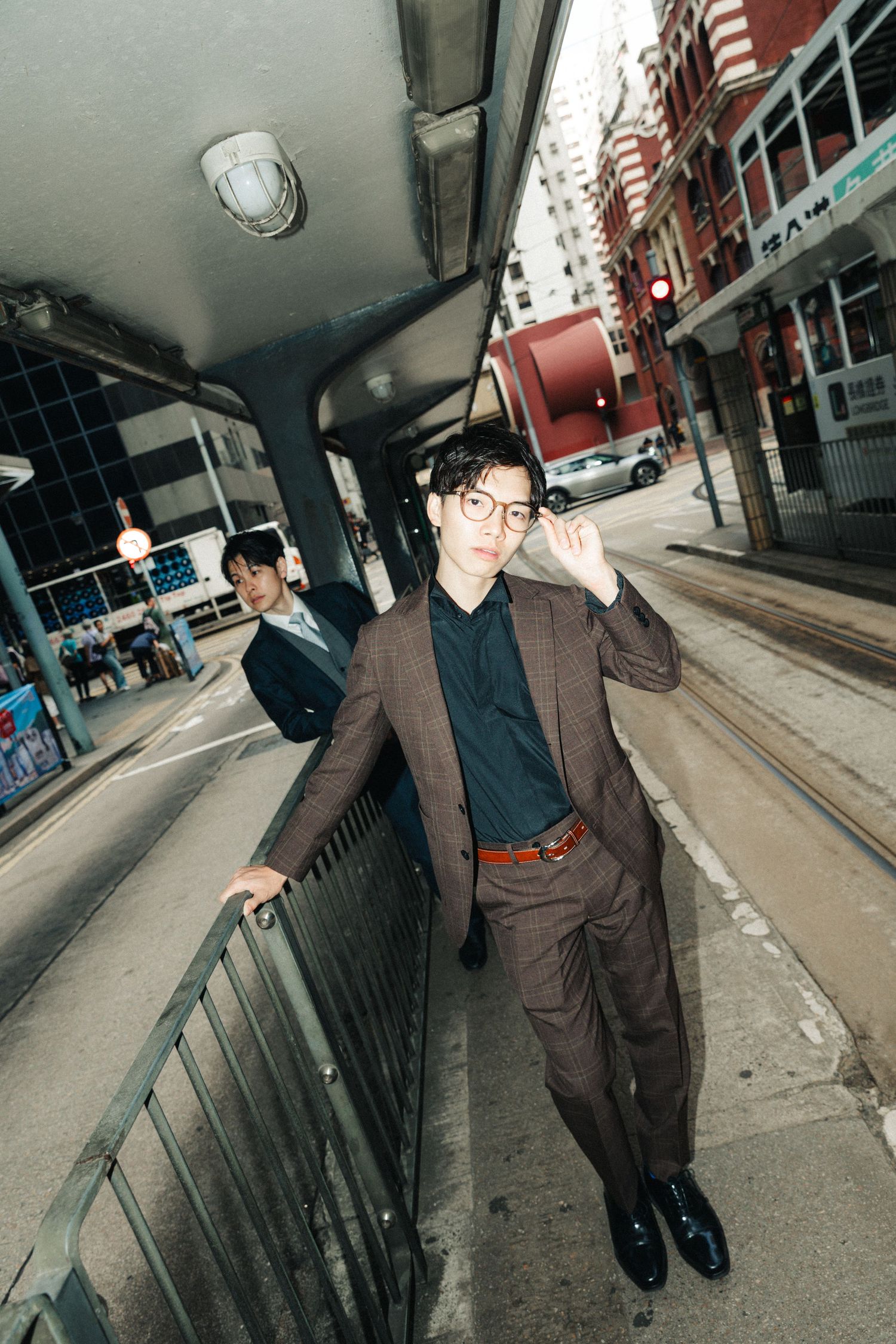 Stylish man in brown suit and glasses leaning on railing in urban Hong Kong street with tram tracks.