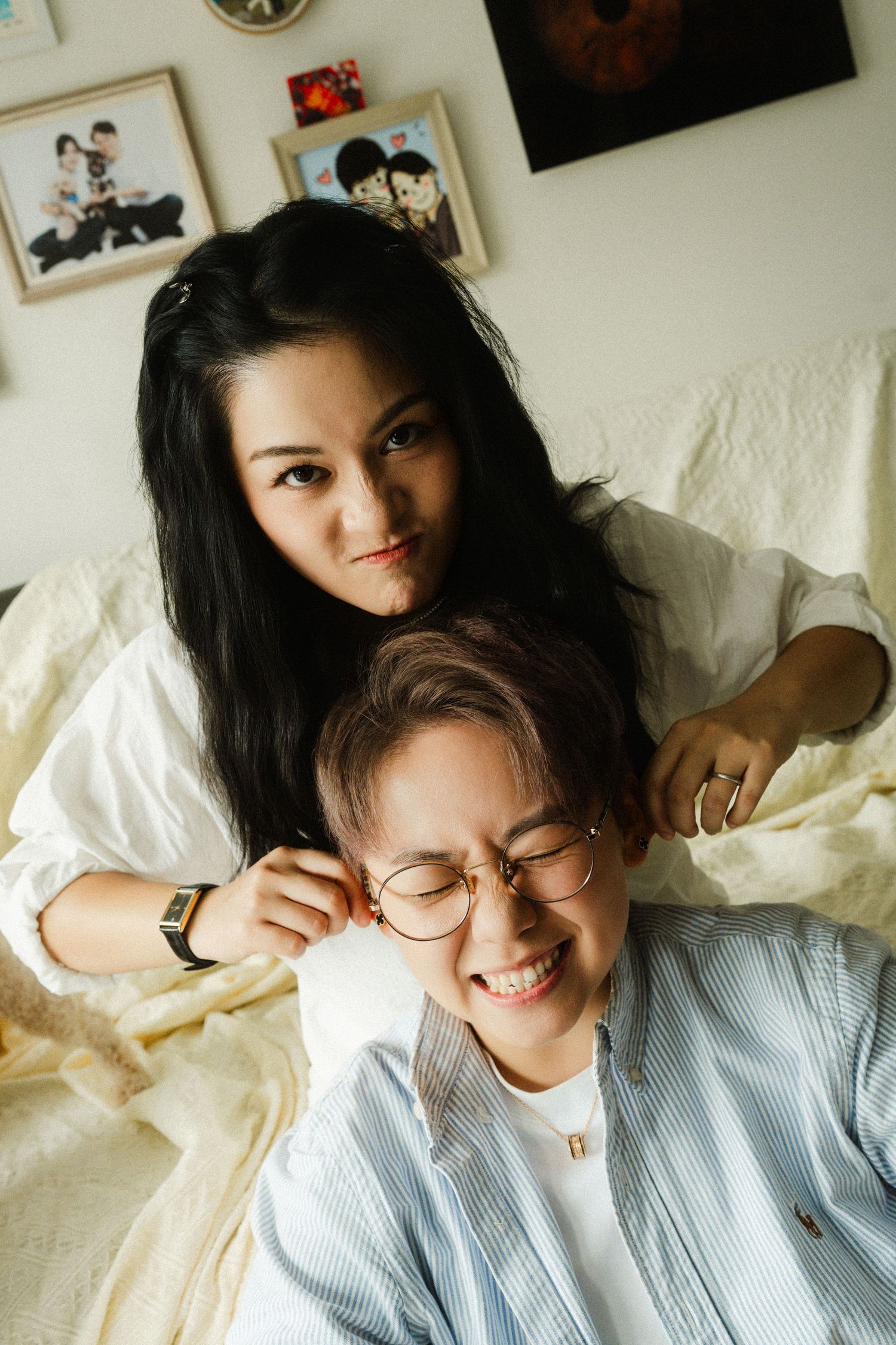 Two women relax on a bed, one playfully touching the other's ear while smiling warmly.