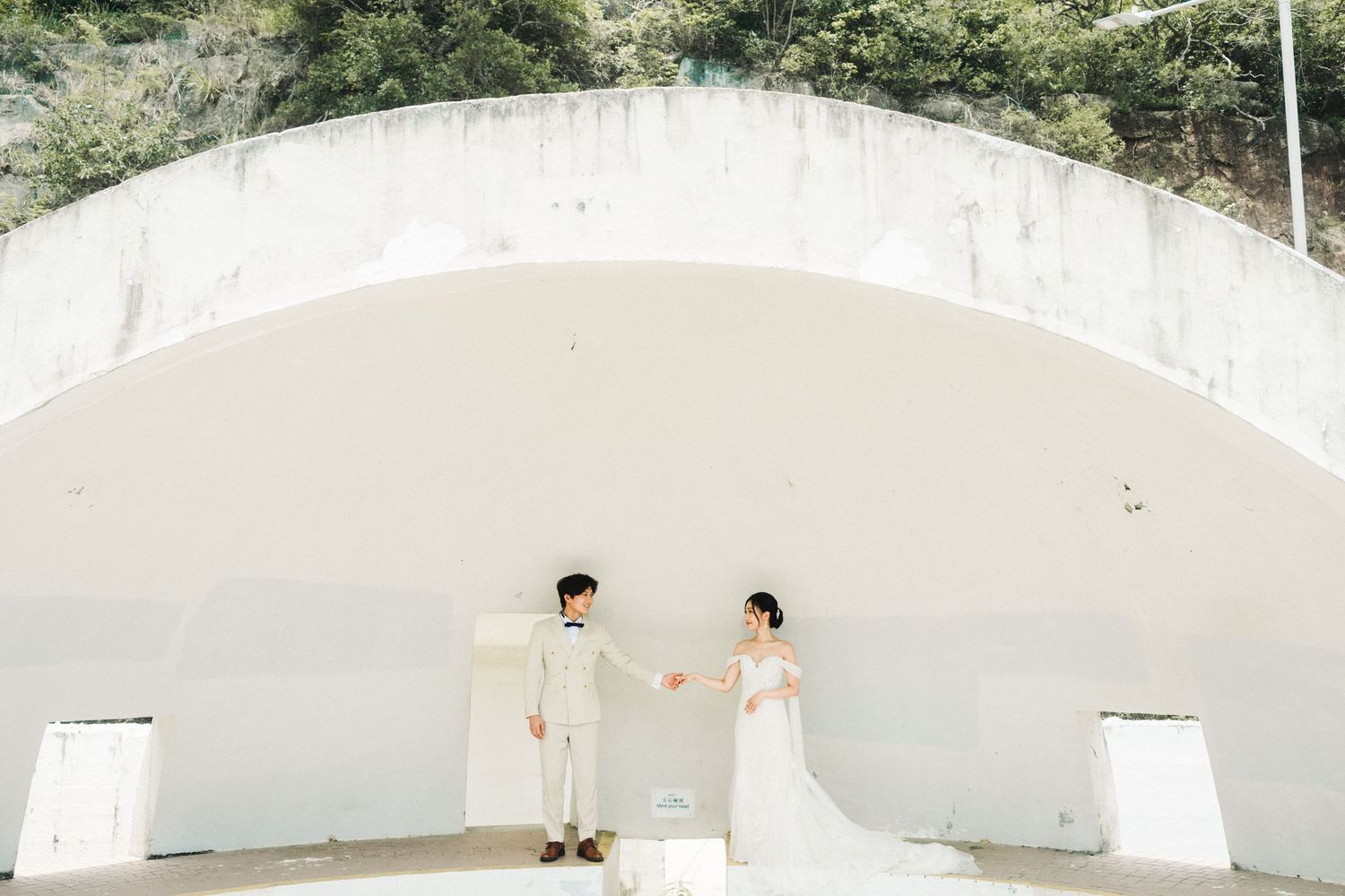 Bride in white gown and groom in beige suit pose under large white arch for wedding photos.