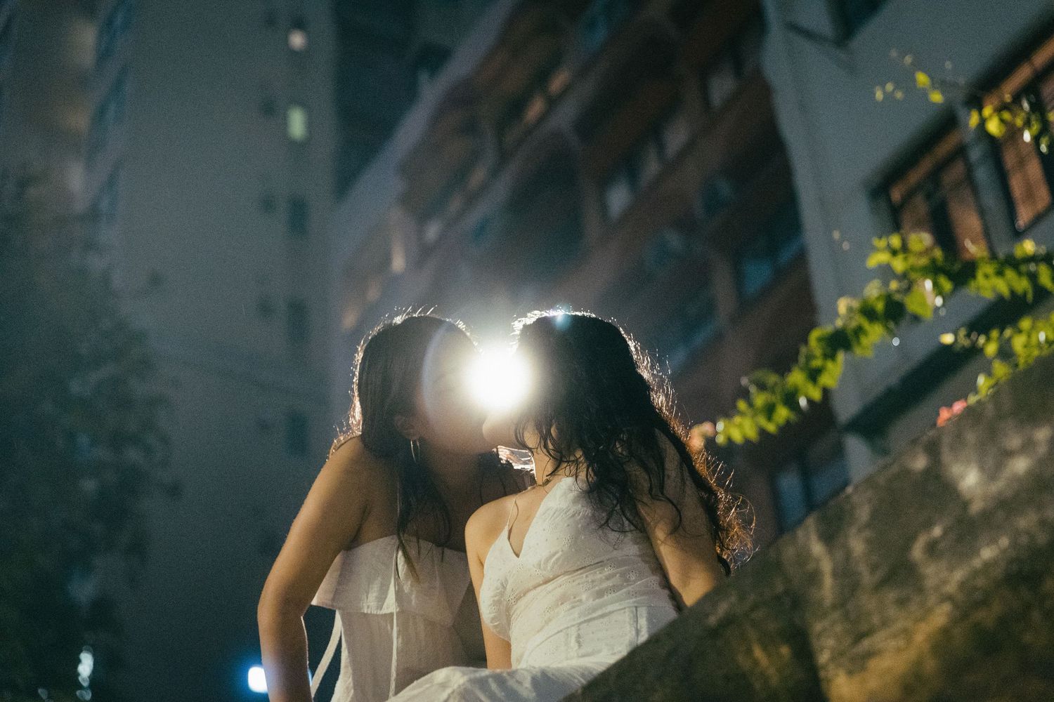 Two women sharing a kiss on a wall at night with city buildings behind them.
