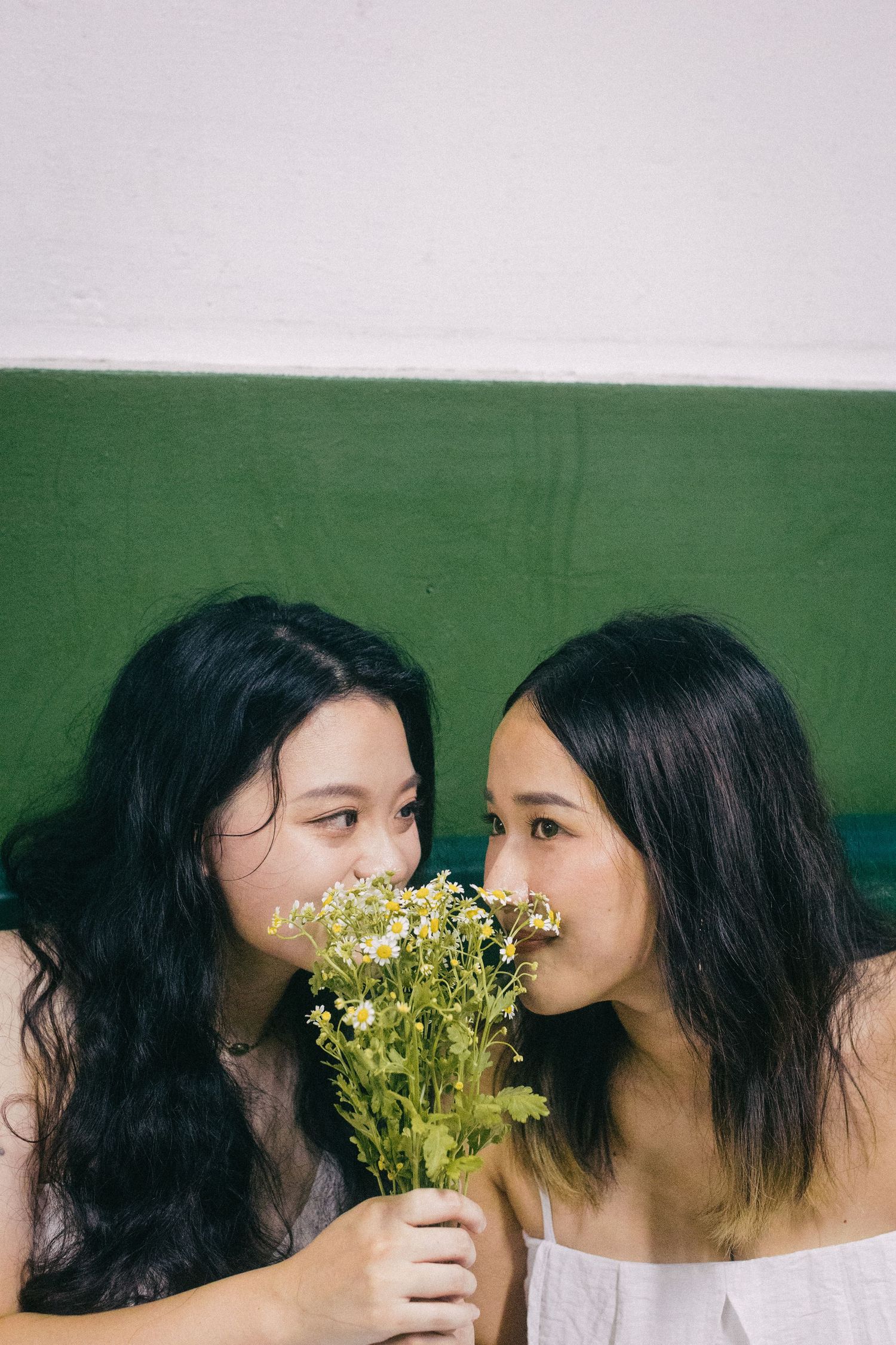 Two young Asian women smelling a bouquet of wildflowers together indoors.