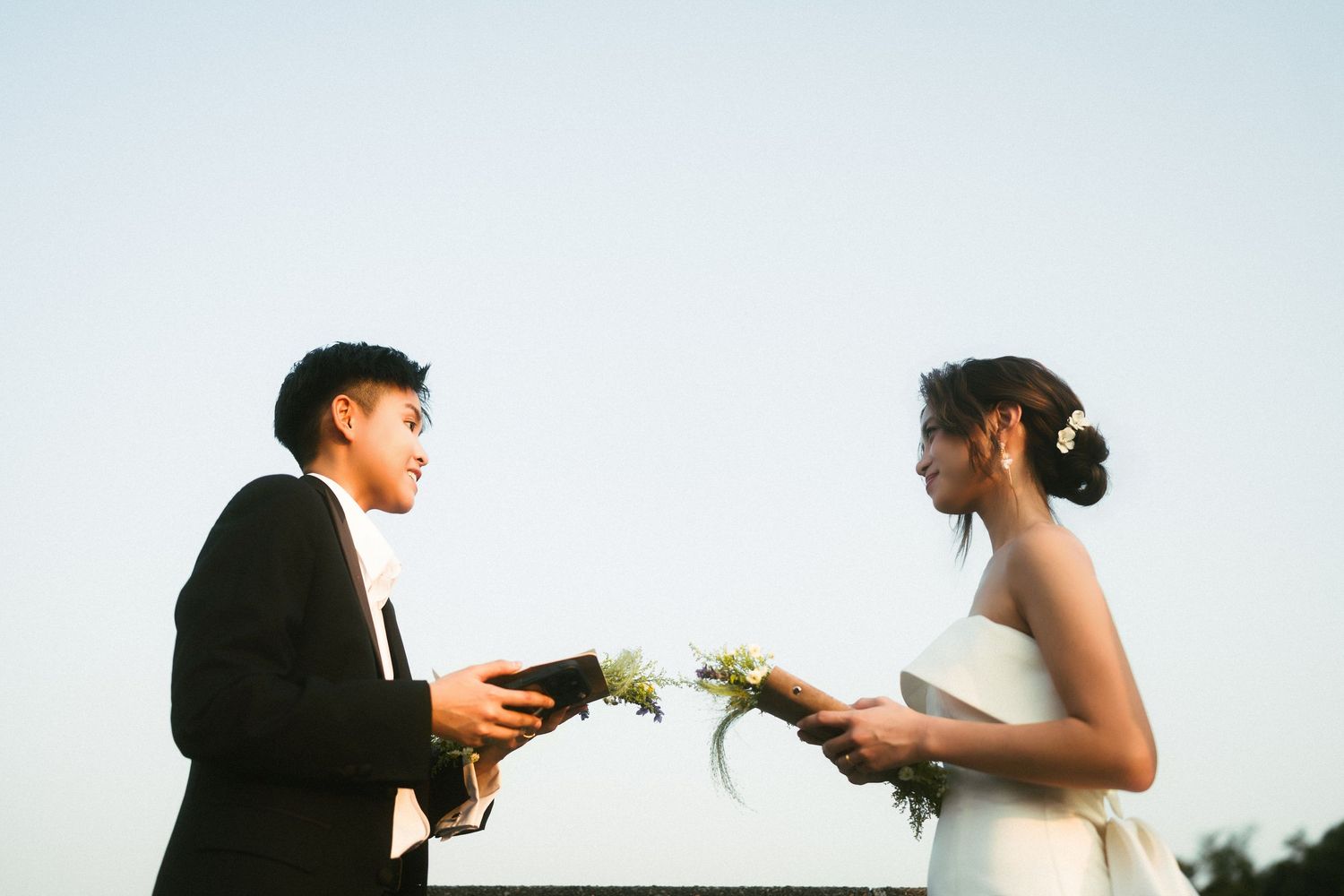 Couple exchanges rings outdoors at dusk, one in a black suit and one in a white wedding dress.