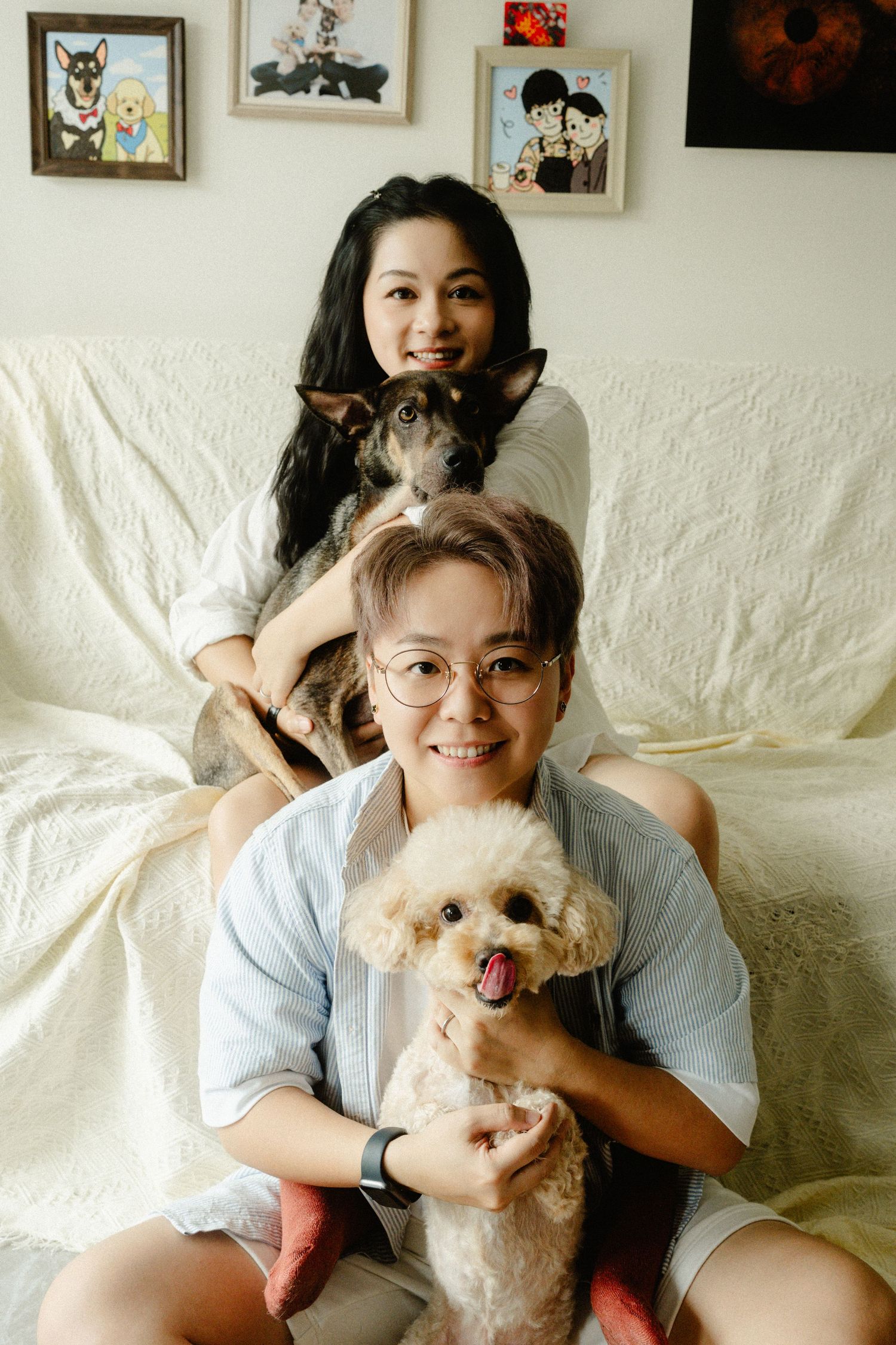 Smiling couple posing on sofa with two dogs, a poodle and mixed breed, with artwork on wall behind.