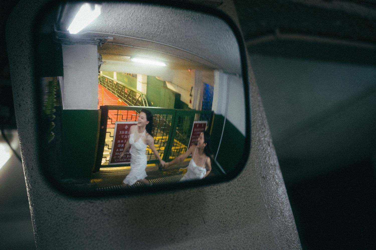 Two women in white dresses reflected in a vehicle side mirror in a parking garage.