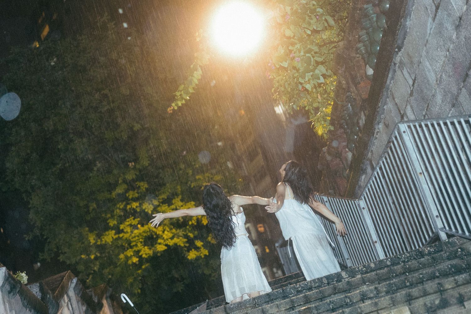 Two women in white dresses hold hands on rain-soaked steps illuminated by warm golden light.