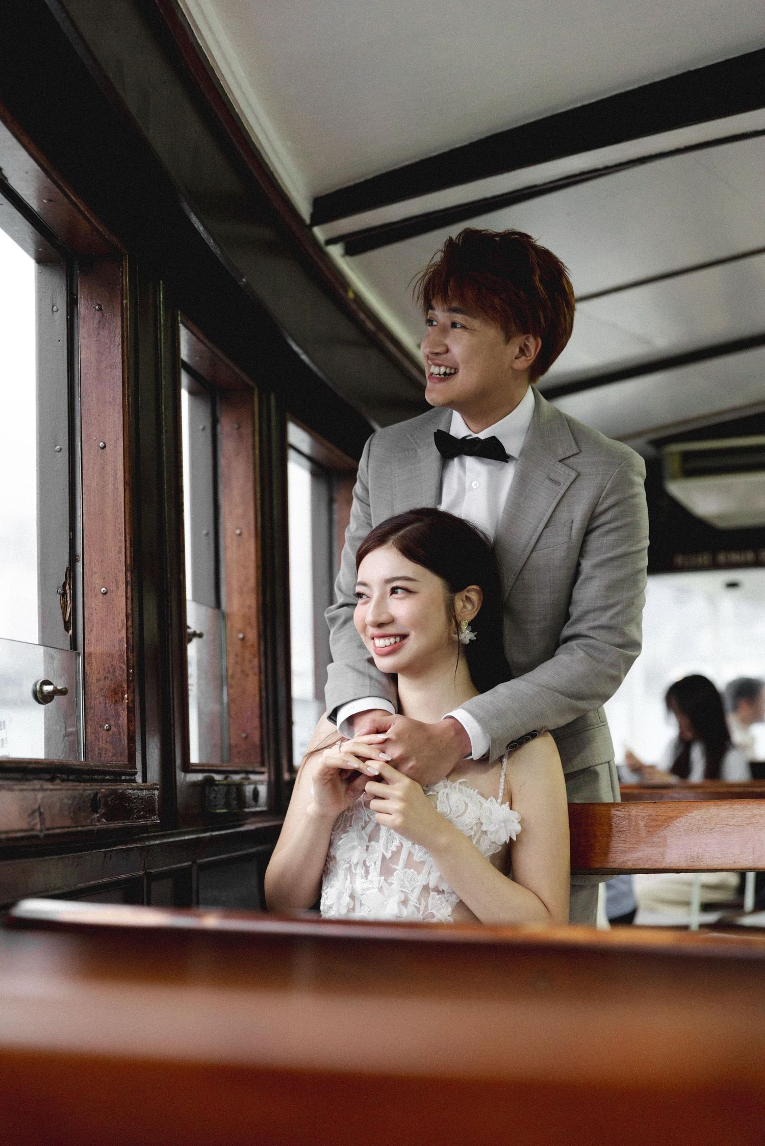 A smiling couple in wedding attire poses on a vintage tram, groom in gray suit embracing seated bride in white lace dress.
