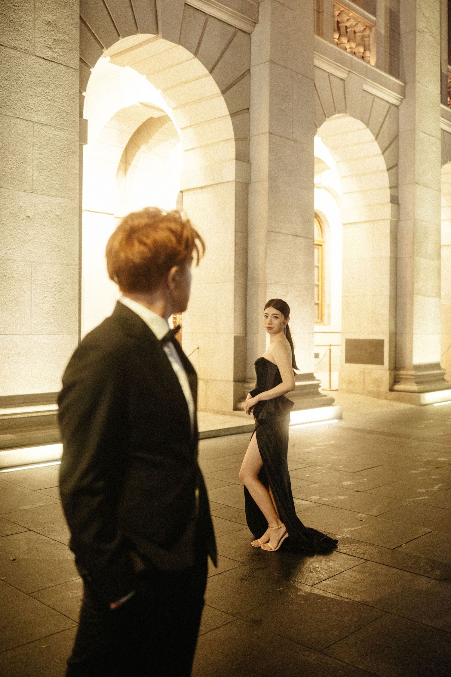 A woman in a black gown glances back at a man in a tuxedo outside an illuminated building.