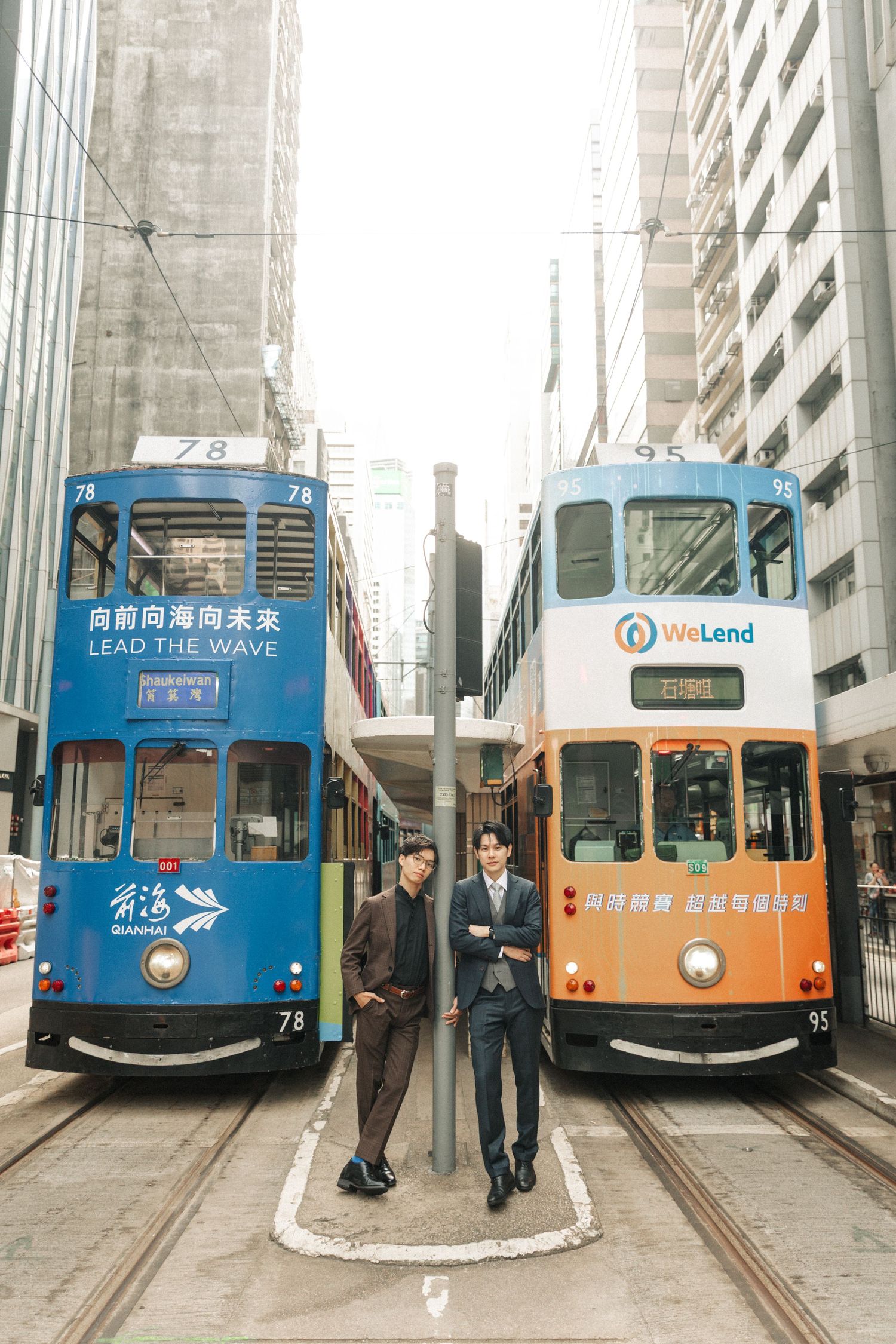 Two suited Asian men stand between a blue and orange double-decker tram in Hong Kong.