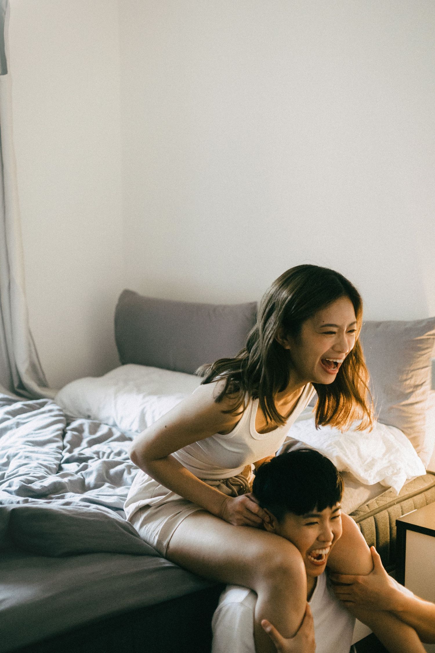 A joyful Asian couple laughing and playing together on a bed in a bright bedroom.