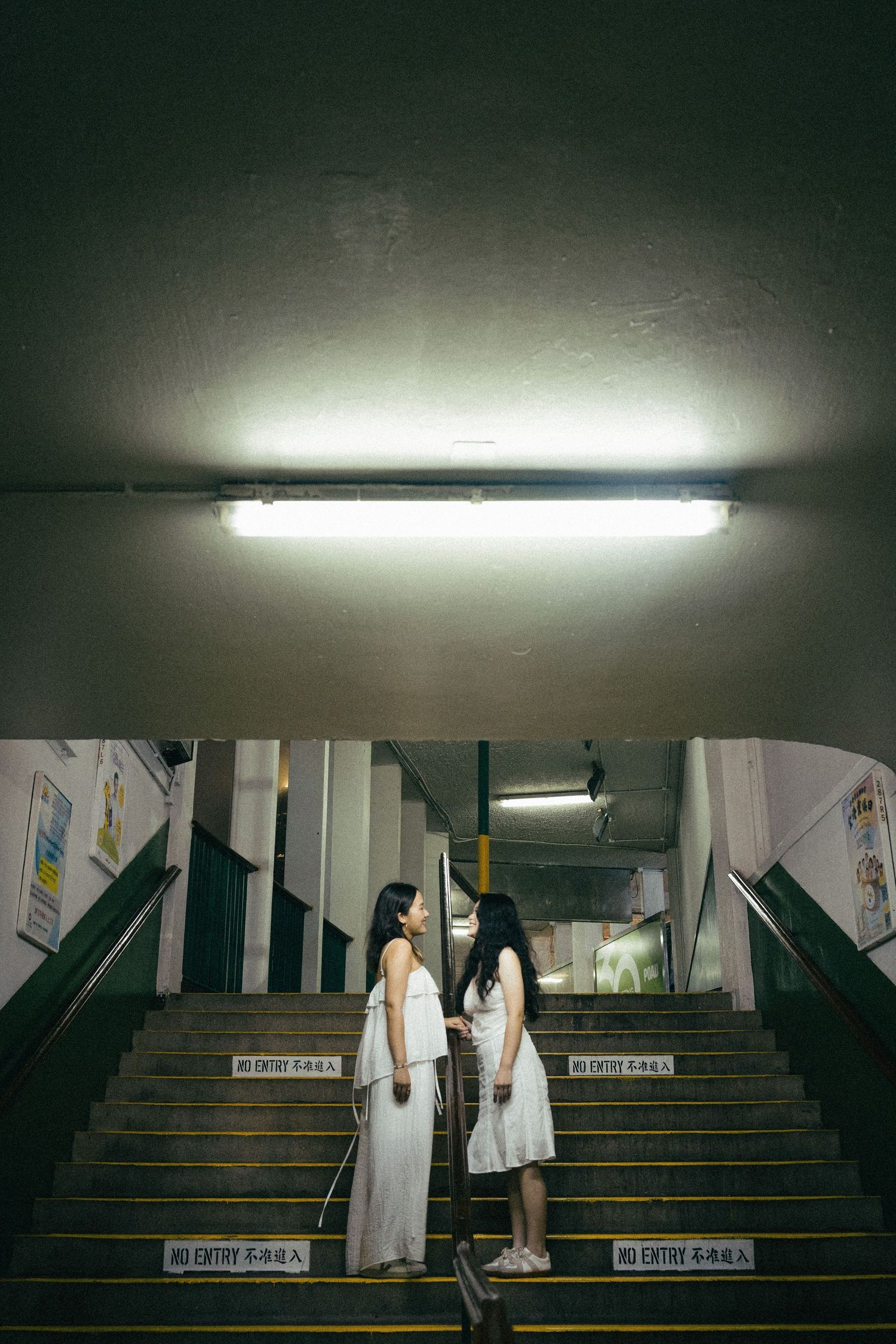 Two women in white dresses stand facing each other on urban stairs under fluorescent light.