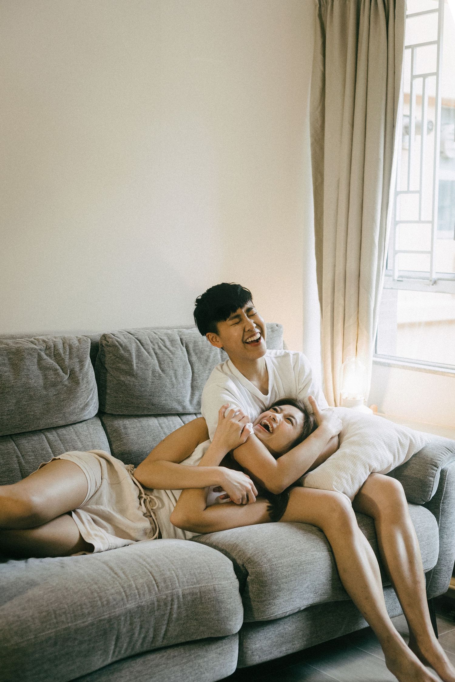 A happy young Asian couple laughing and cuddling together on a gray sofa in a sunlit living room.