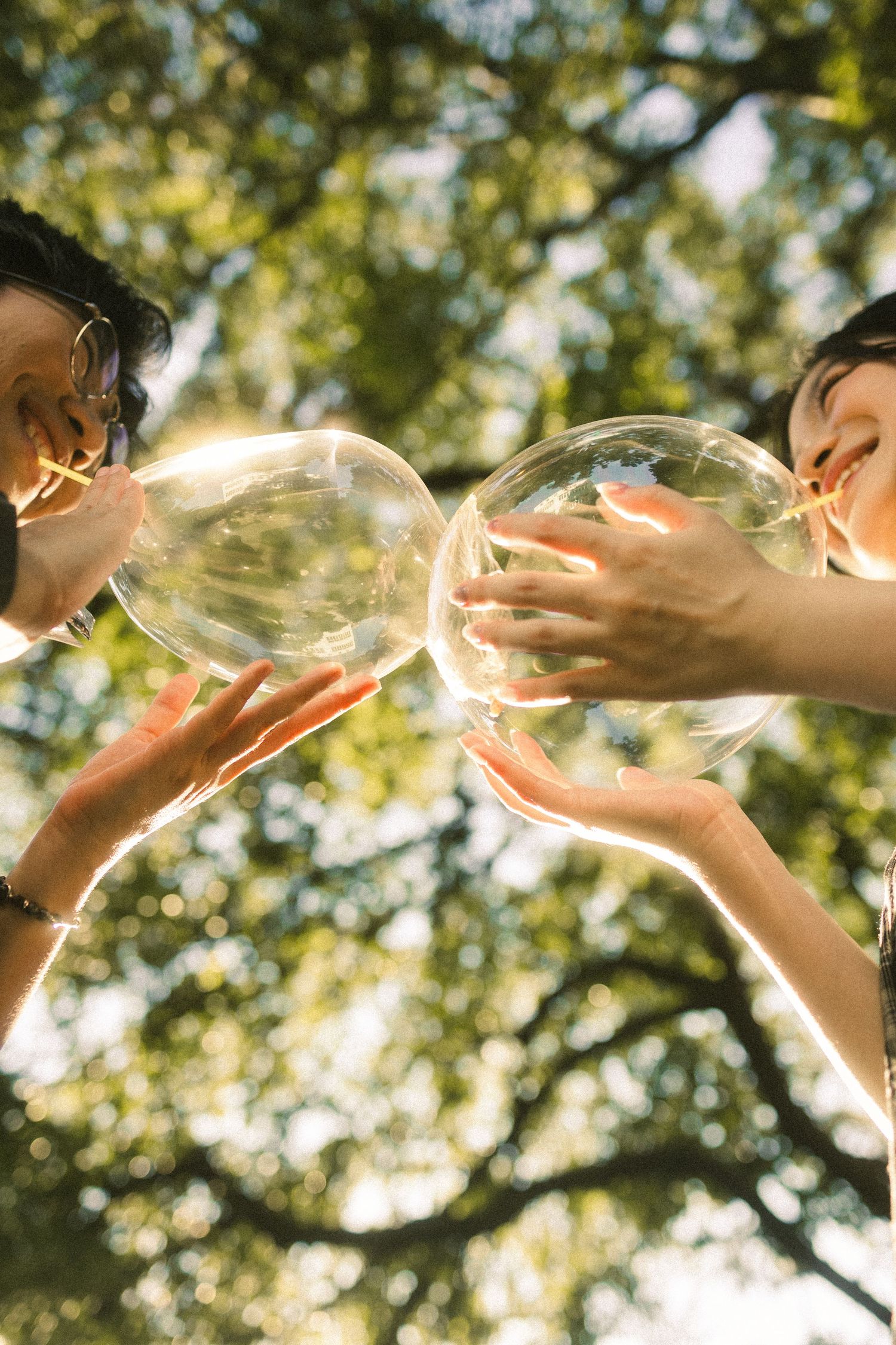Low angle view of two people touching large transparent bubbles together under green trees.