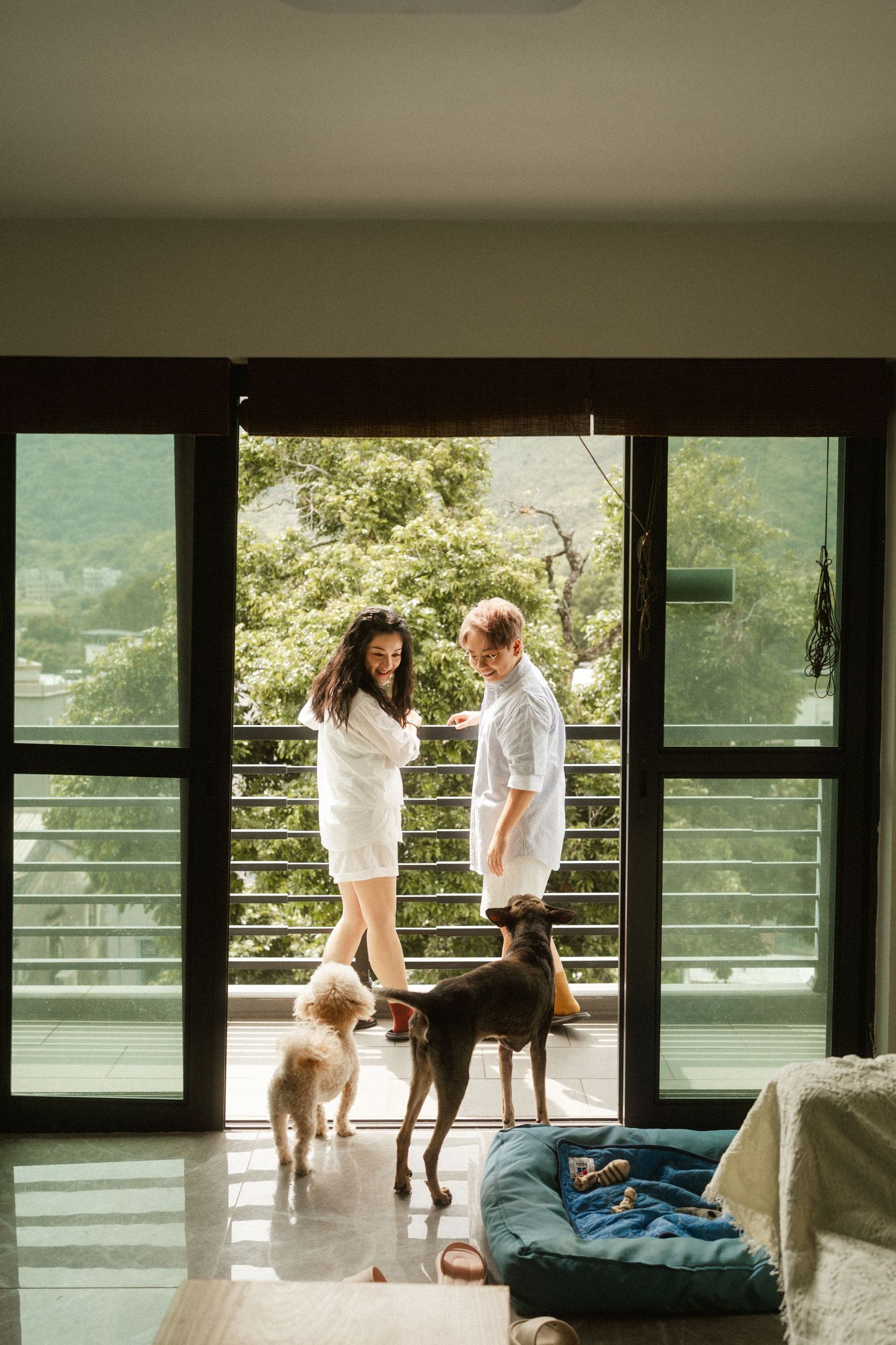 Couple in white outfits standing on balcony with two dogs and lush green mountain view.