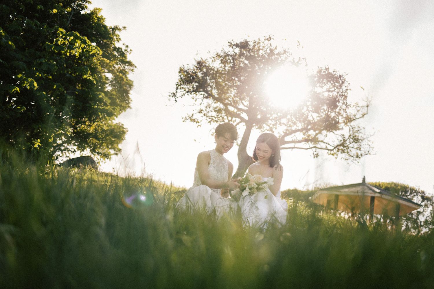 Two brides in white gowns sit together in lush green grass under a sunlit tree.