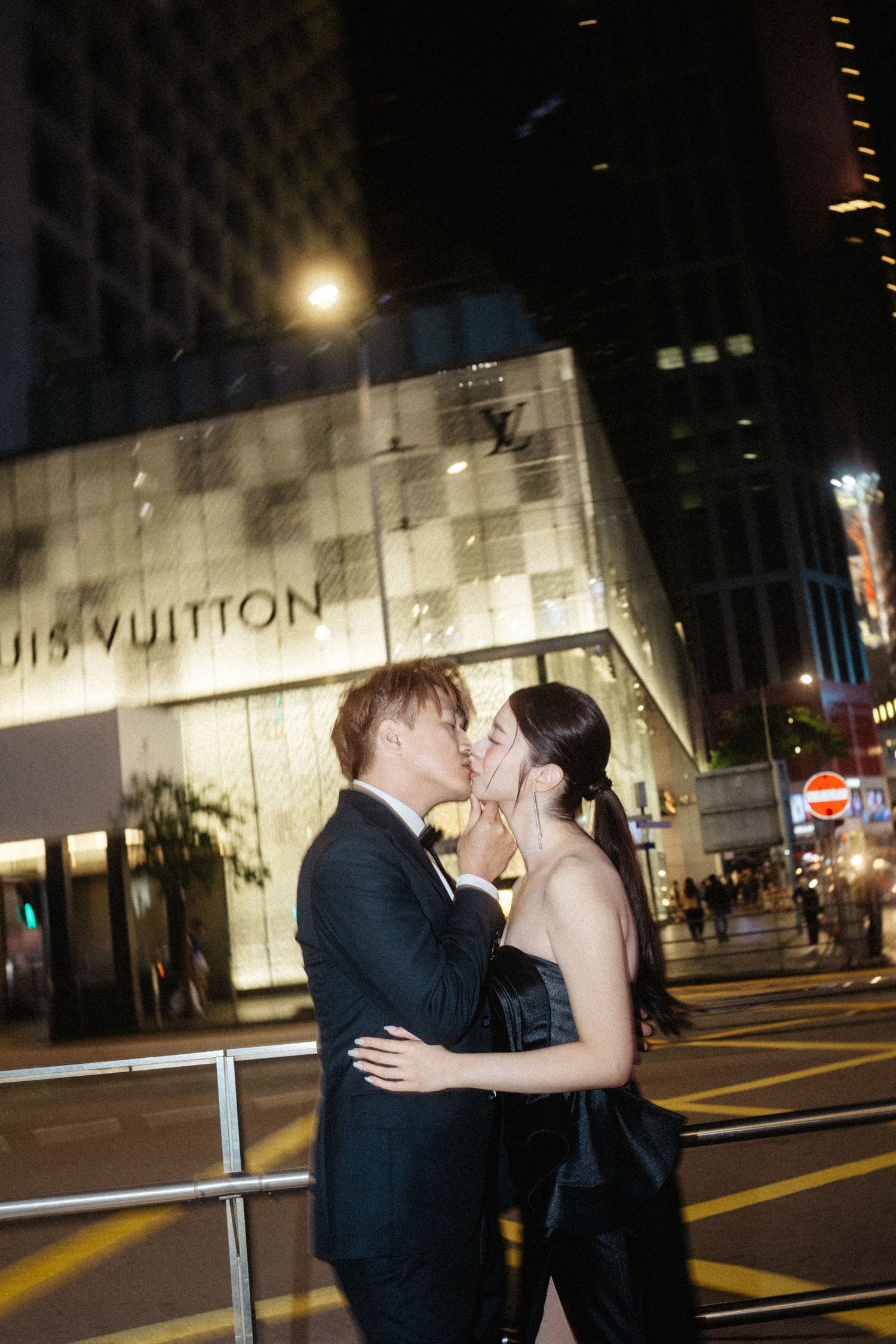 A couple in black formal attire kisses on a city street at night in front of a Louis Vuitton store.