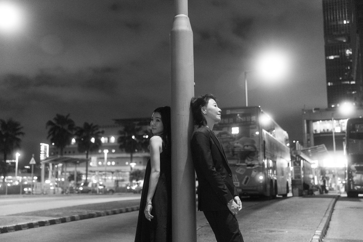 Black and white photo of two people leaning against a lamppost on a city street at night.