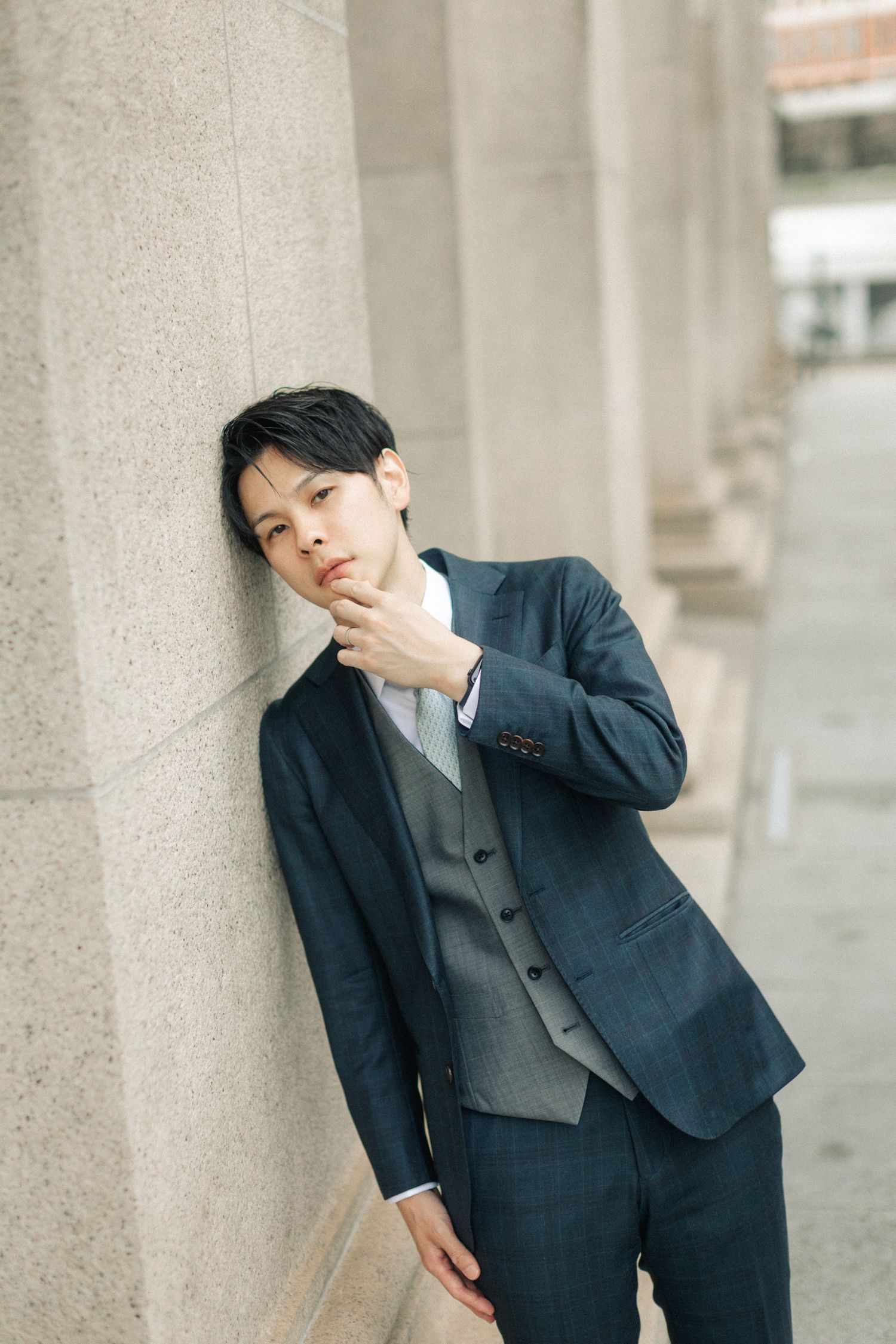 A young Asian man in a navy three-piece suit leans against a stone column thoughtfully.