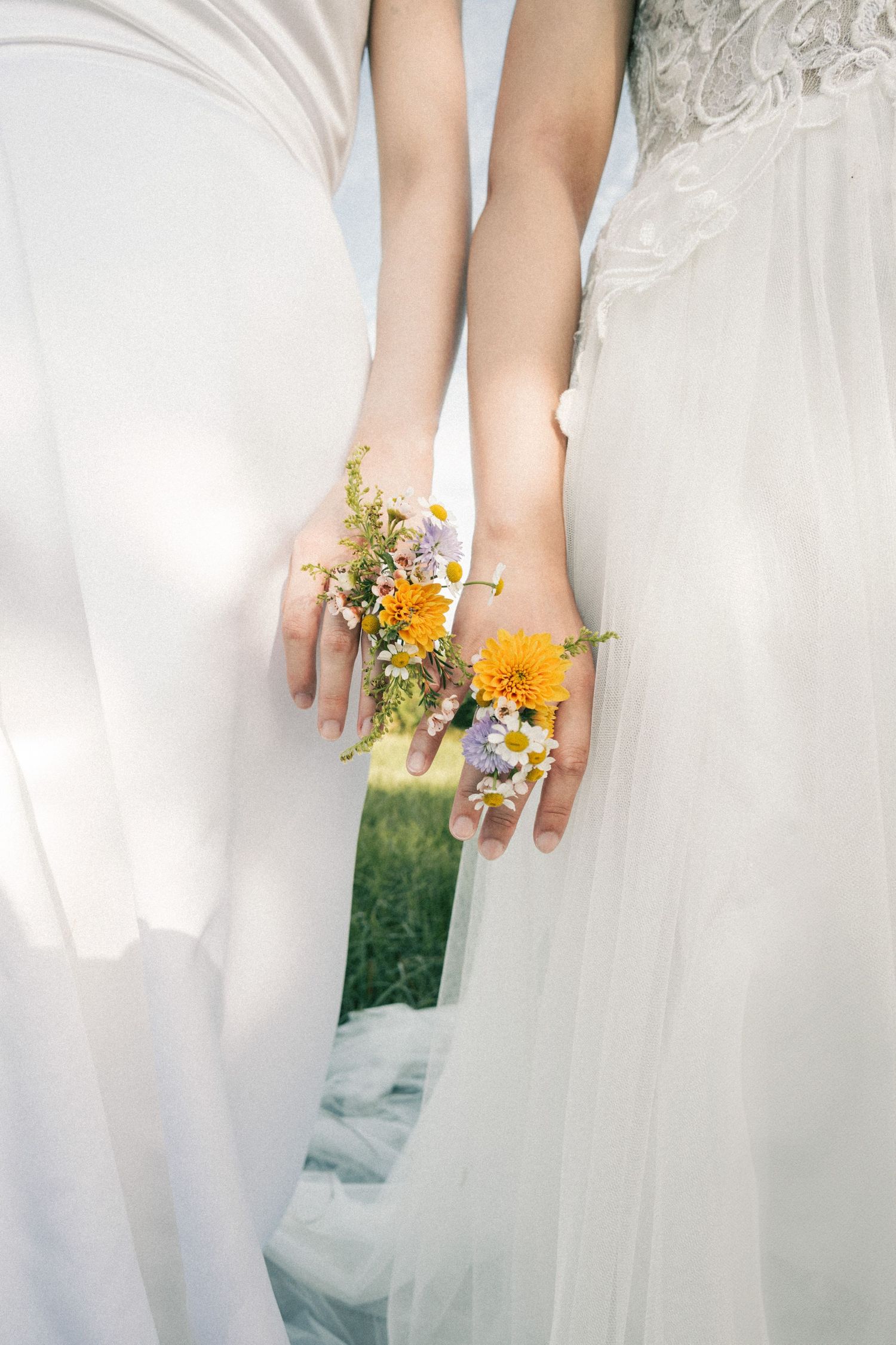 Two brides stand side by side in white gowns, hands intertwined holding colorful wildflowers with a mirror reflection.