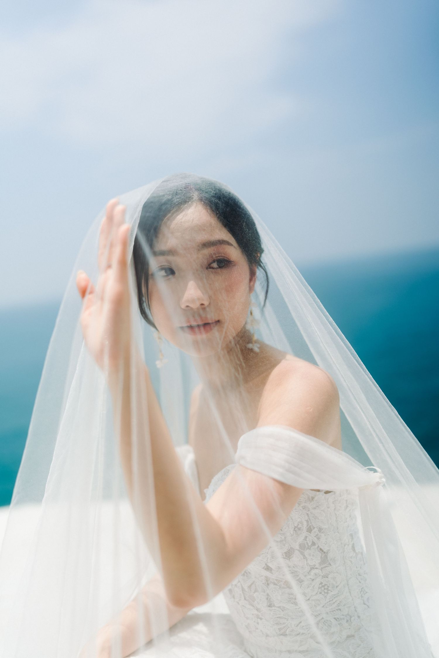 Asian bride in lace off-shoulder gown gazes through sheer veil with blue ocean in background.