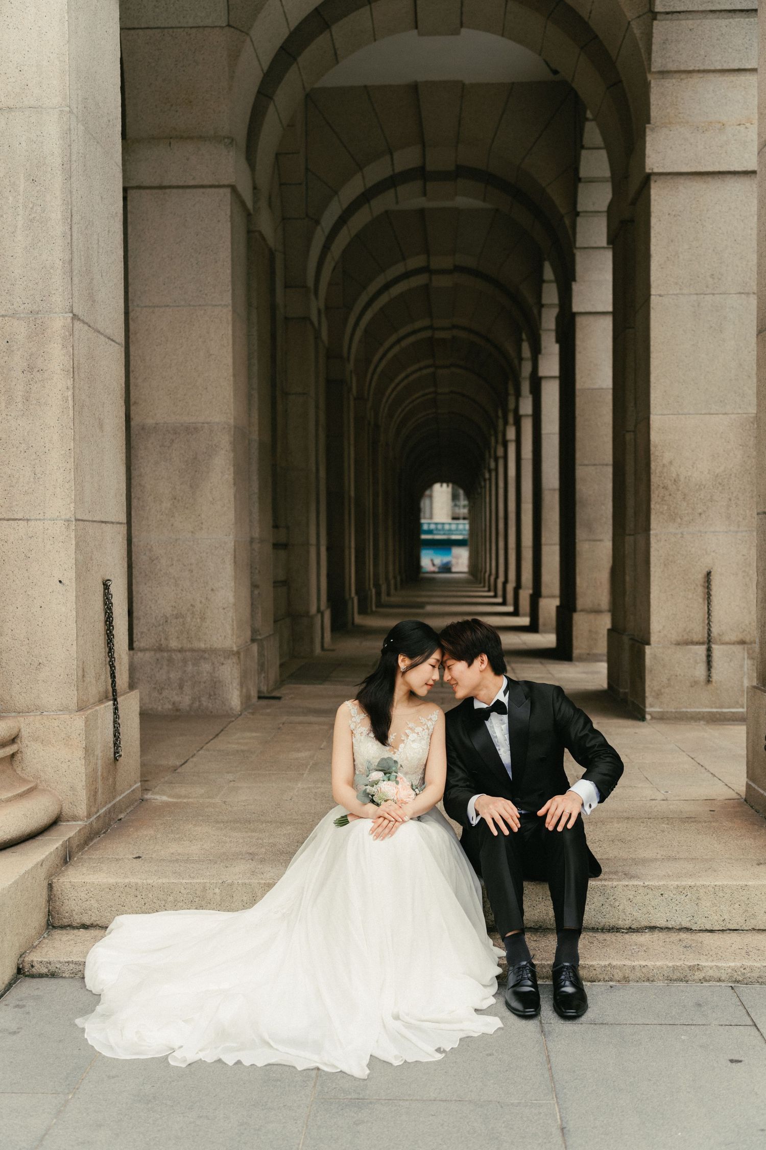 Bride and groom sharing a tender moment seated in an elegant arched stone hallway.